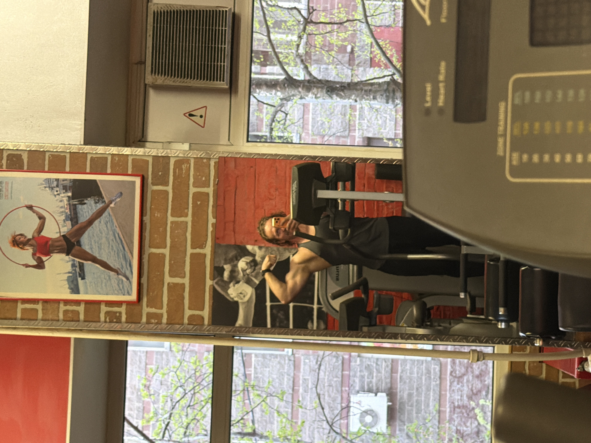 A medium indoor shot shows a gym facility in St. Petersburg, Russia. The background includes brick walls with a poster of a gymnast in red attire with a hoop, and artwork. A person is reflected in the mirror, visible with a flexed bicep. A window shows a building and tree branches outside. The foreground features a workout machine console.