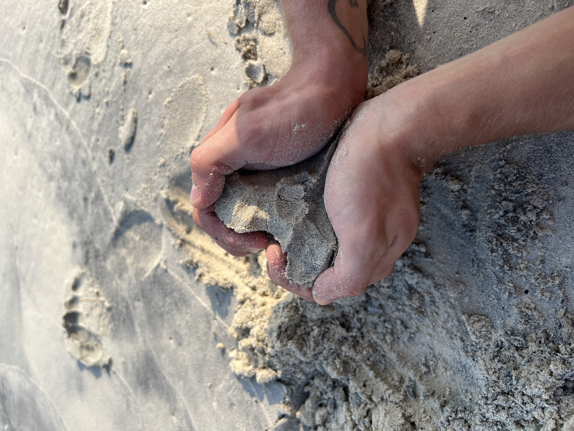 A close-up photograph depicts two hands holding a clod of wet sand. The sand is a mixture of light and dark grains. The hands are light-skinned and covered in fine grains of sand. The left hand has a tattoo on the inner wrist area. The background shows more sand, with varying textures and moisture levels and a footprint is visible on the left side. Based on the sand type, lighting, and presence of footprints, it's likely the picture was taken on a beach, possibly in the city of Nha Trang, Vietnam, given the user's prompt. The action is hands holding sand.
