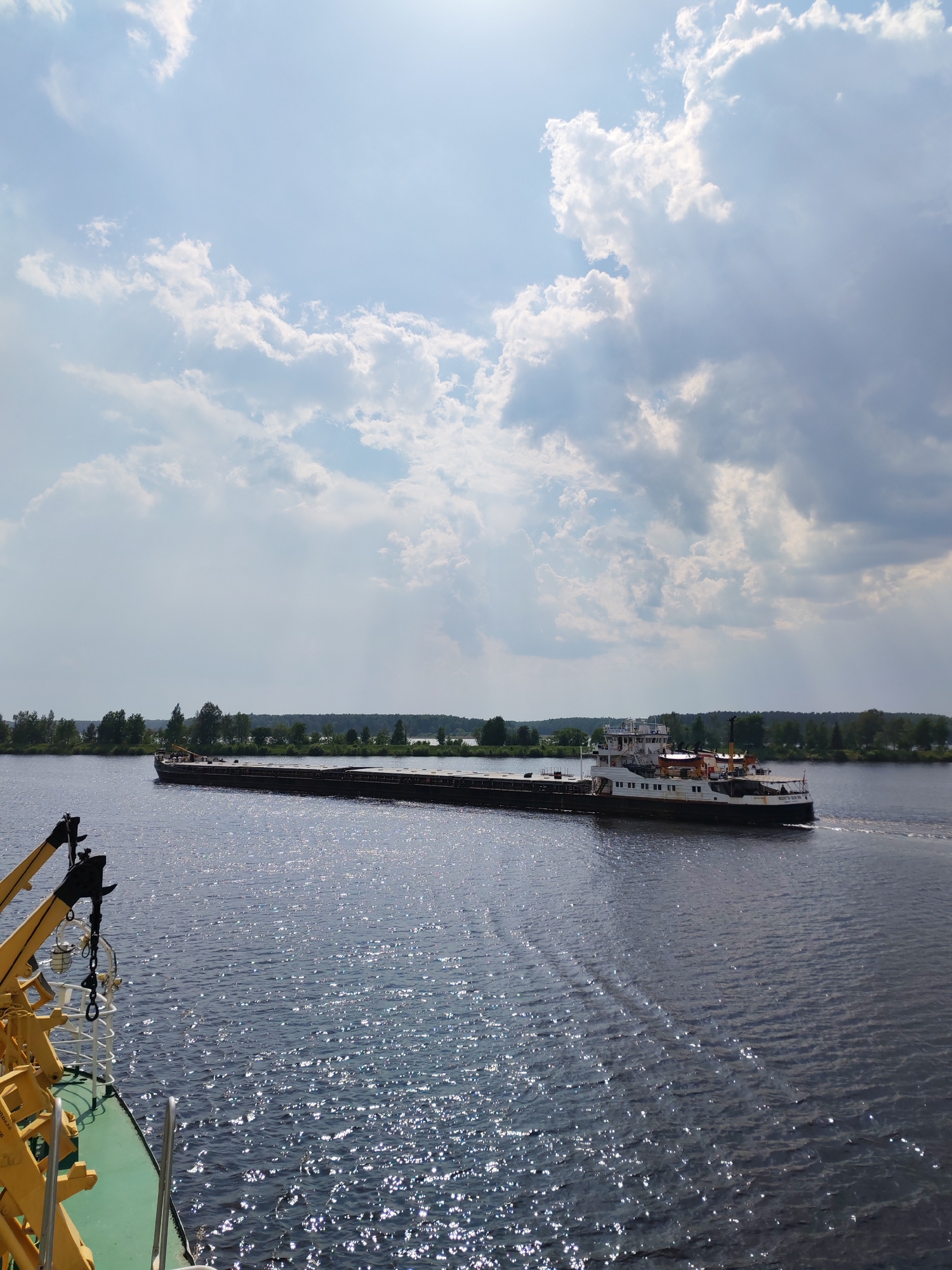 A wide shot captures a barge moving along a body of water on a partly cloudy day. The sky is filled with a mix of white and gray clouds, with a bright sun shining through. The water reflects the sunlight, creating a shimmering effect. The barge is long and flat, with a tugboat or other propulsion unit attached to the rear. The shoreline is visible in the distance, lined with trees and vegetation. The barge appears to be in motion, leaving a wake behind it. A small piece of a boat with yellow parts is visible in the bottom left corner of the shot. The location is Unknown, Unknown.
