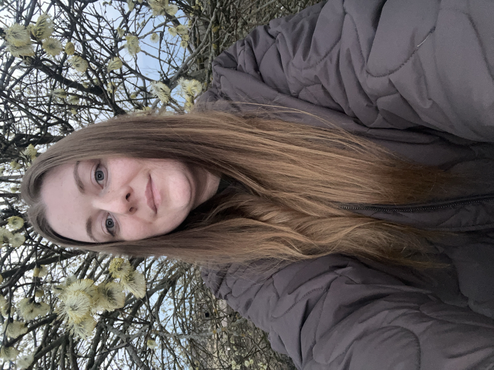 A fair-skinned woman with long brown hair is centered in the image. She is wearing a puffy gray coat, zipped up to the neck. Behind her, there are blossoming trees with fuzzy, yellow-white flowers and bare branches reaching up towards a pale sky. The picture may have been taken outdoors in Razdolye, Russia, though there is no specific cityscape or signage to confirm the location.
