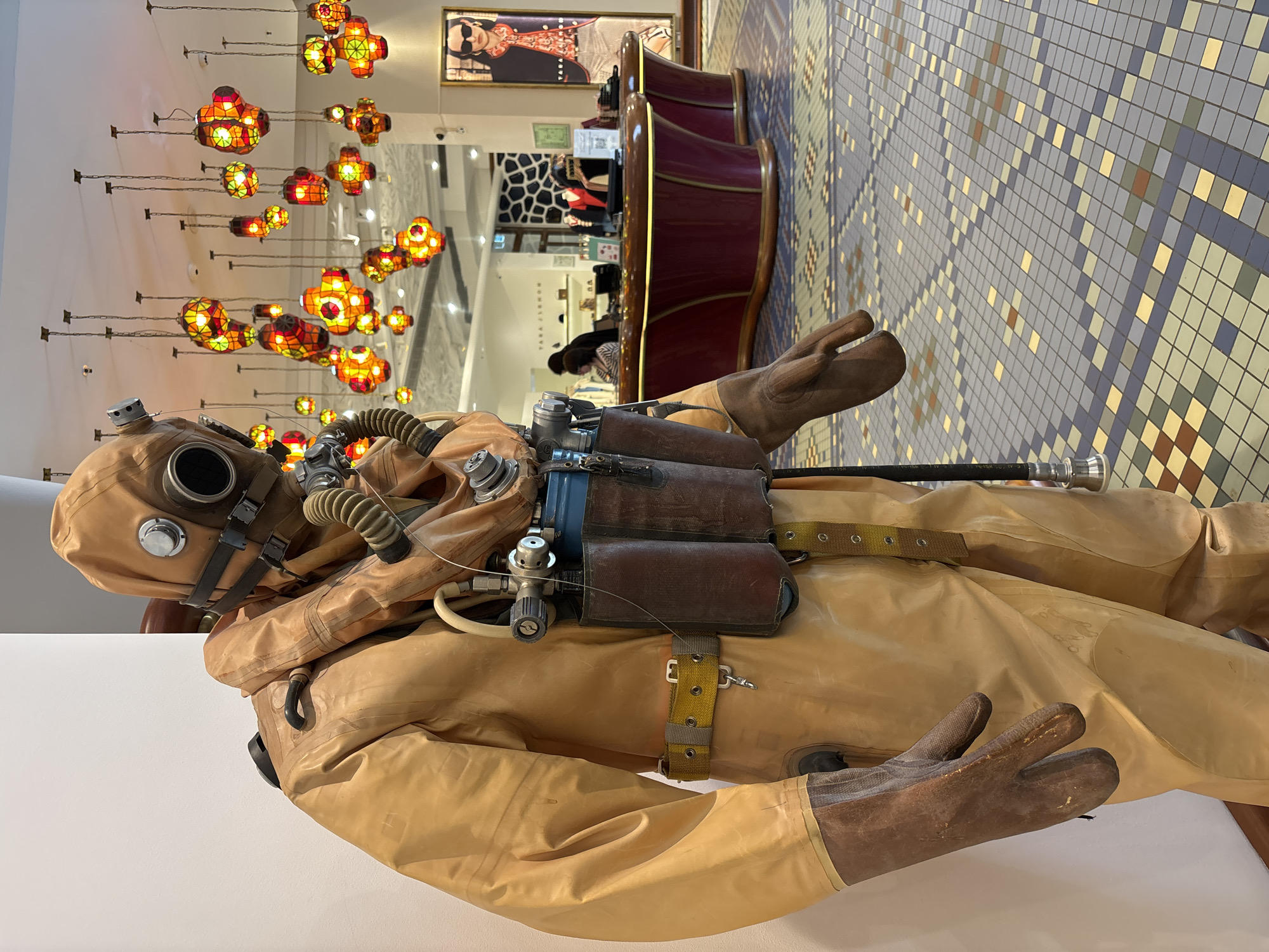 A vintage deep-sea diving suit is displayed in what appears to be a retail setting. The suit is predominantly tan/brown, and includes a helmet with a circular viewport and hoses, brown gloves attached to the sleeves, and canisters strapped to the chest. The suit is positioned against a white wall. The background features a mosaic tile floor, colorful hanging lights, and retail displays including counters and mannequins. The scene is likely within a retail establishment in Saint Petersburg, Russia.