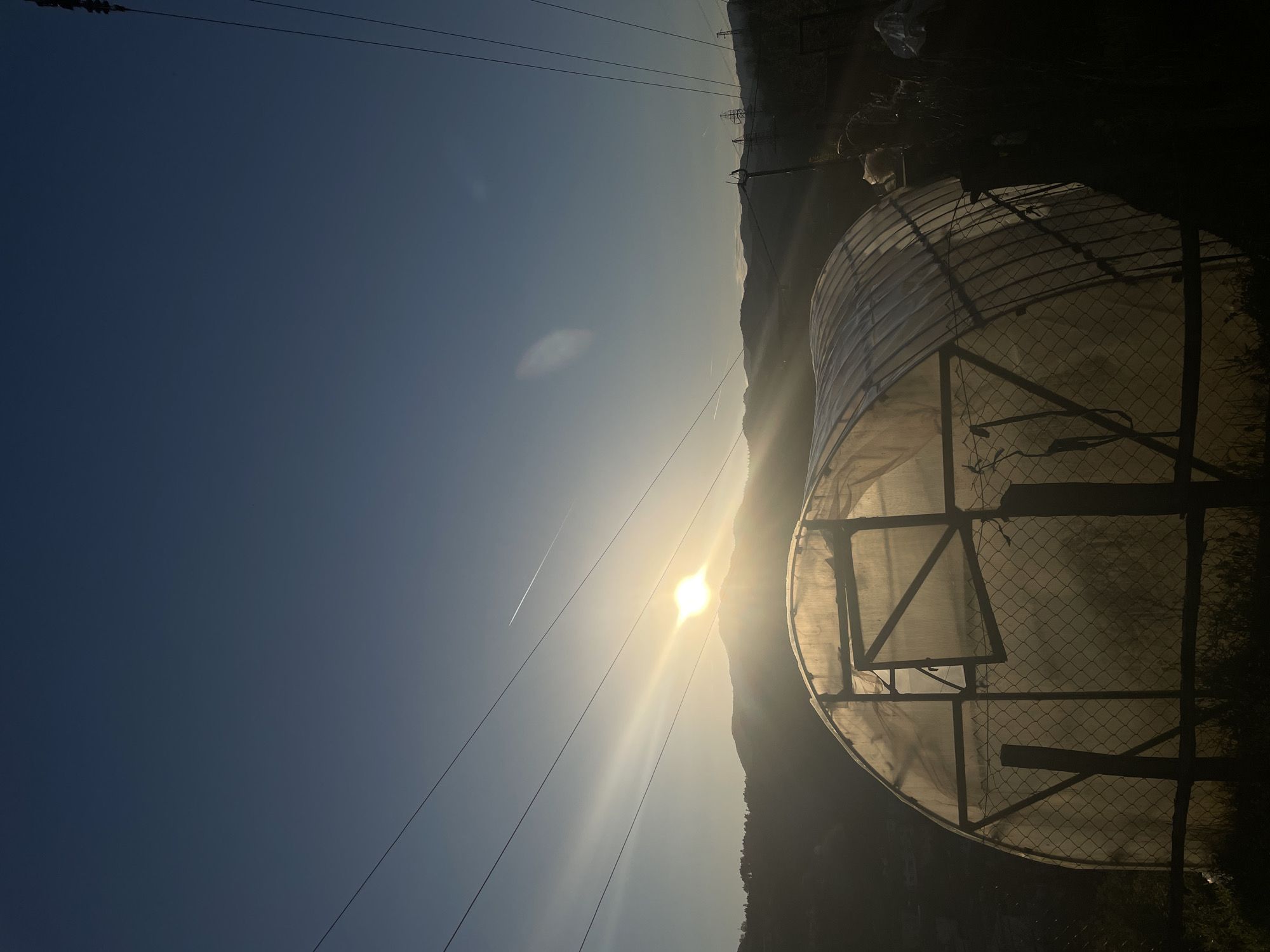 The image shows a partially obstructed view of the sun behind a mountain ridge with power lines extending from the top left corner towards the mountain. To the right, there is a partially visible domed structure possibly covered in translucent plastic or a similar material, surrounded by a wire fence. The image appears to have been taken in an area with mountainous terrain, which could be consistent with the geographical location of Zlatograd, Bulgaria. No people are visible in the scene. There is a contrail from an airplane across the sky.