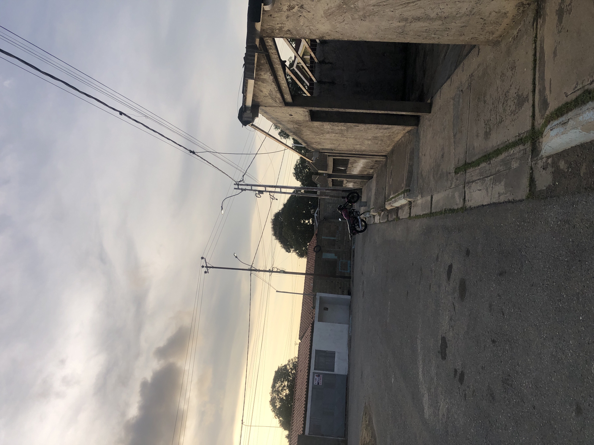 This is a street scene in Altagracia, Venezuela. The image, oriented sideways, shows a narrow asphalt road lined by buildings on either side. The structures are predominantly concrete, with a mix of finished and unfinished construction. Overhead, a network of power lines crisscrosses the sky. A motorcycle is parked on the side of the road. The sky is overcast, with an orange glow hinting at either sunrise or sunset. There are no people visible in the frame.