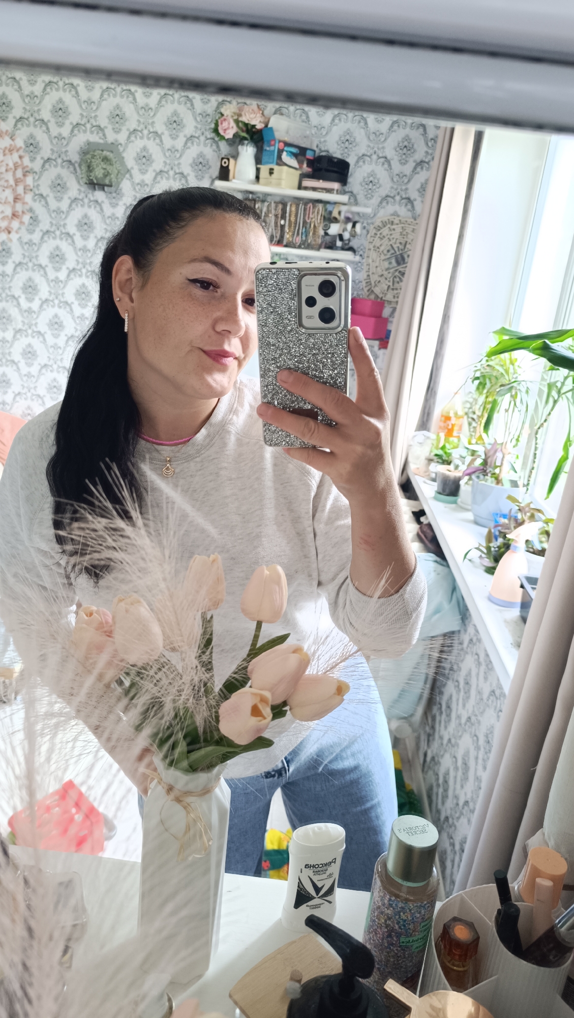 A woman is taking a selfie in a mirror. She is wearing a light gray sweatshirt and blue jeans. She has long dark hair pulled back in a ponytail and is wearing a pink necklace and small hoop earrings. In her arms, she holds a white vase with a bouquet of pale pink tulips and feathery grass. The background shows a wall with patterned wallpaper, shelves filled with various items including decorative objects, jewelry, and what appear to be cosmetics. A window with curtains is visible to the right, with several potted plants on the windowsill. The scene appears to be in a residential interior, likely a bedroom or dressing area, in Chaikovsky, Russia, as indicated by the presence of Russian text on a deodorant stick in the foreground.