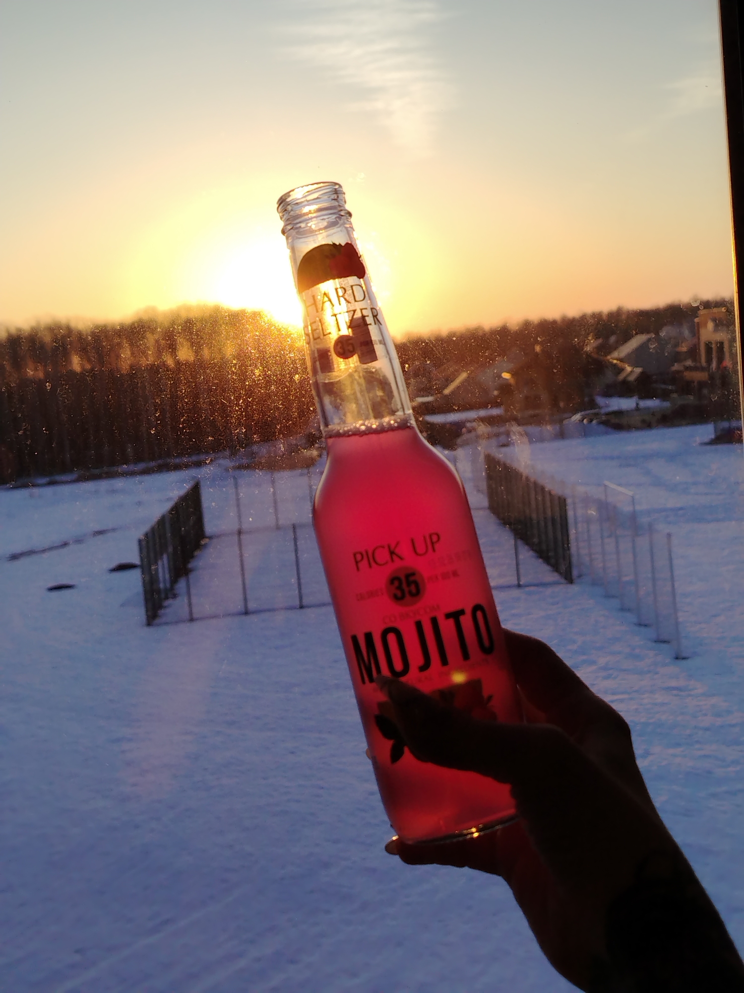A hand holds a bottle of "Pick Up Mojito" hard seltzer in front of a window with a snowy landscape and a setting sun. The bottle is clear with a pink liquid inside and has "Hard Seltzer" and "Mojito" printed on it. Outside the window, the ground is covered in snow, and there are rows of short fences. In the distance, there are trees and buildings. The sun is setting on the horizon, casting a warm glow over the scene. The scene is in Noviy Urengoy, Russia.