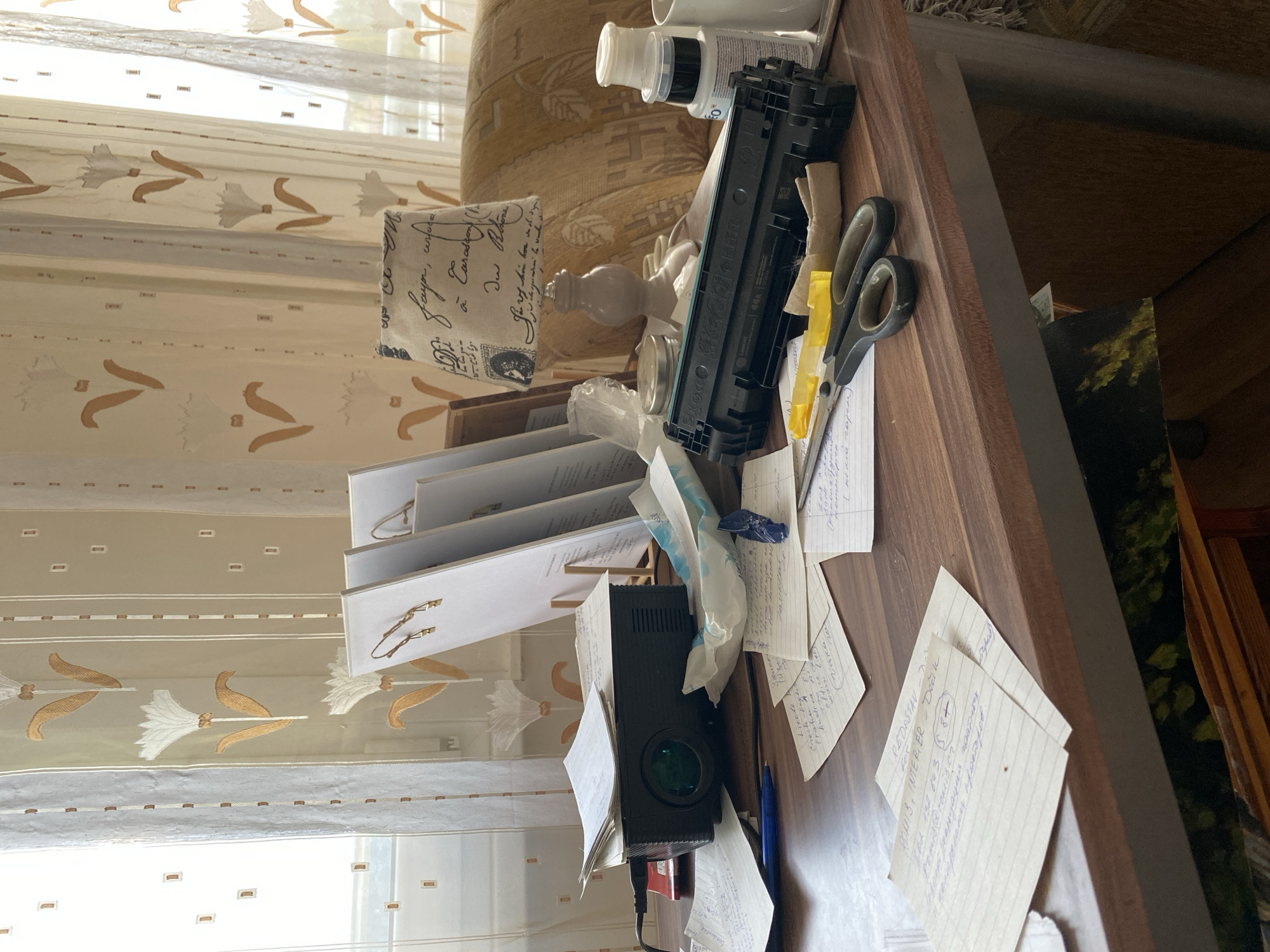 The image depicts a cluttered wooden desk in an indoor setting. The desk is covered with various items, including papers with handwritten notes, a black projector, a printer toner cartridge, scissors, a blue pen, a pack of wet wipes, and several bottles. Behind the desk, a patterned curtain with floral motifs is visible, and a portion of a couch or armchair is also present. The lighting suggests it is daytime, with sunlight filtering in from a window. The scene appears to be a personal workspace, possibly a home office. The location is Bílina, Czechia.