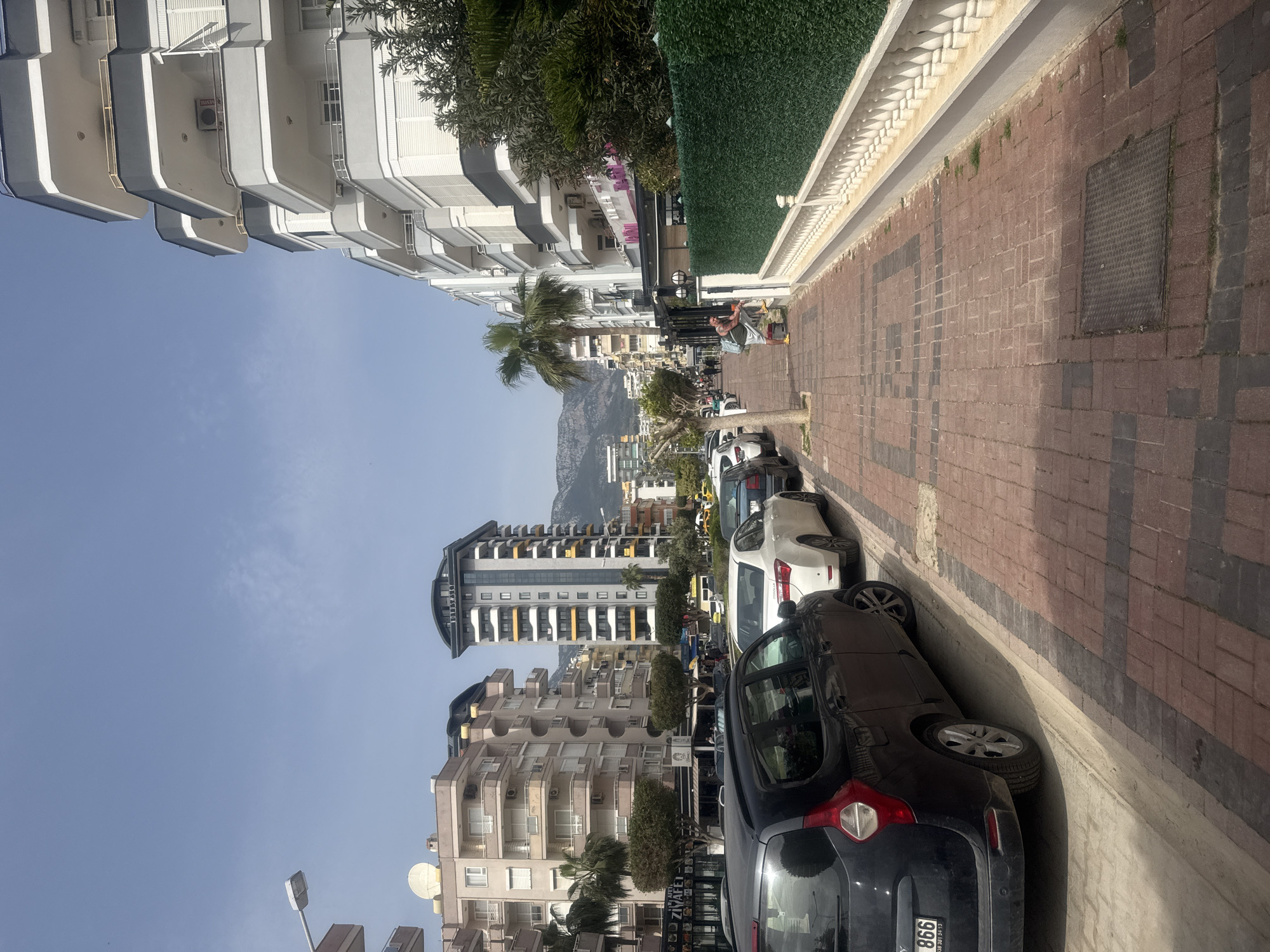 The image depicts a street scene in an urban area. On the left side of the frame, there are tall, white buildings with multiple balconies. Parallel to the buildings runs a pedestrian walkway paved with red bricks with a patterned border. A row of cars is parked along the curb. In the background, a mountain is visible against a clear sky. The scene appears to be outdoors during daylight. A person is standing near a green barrier in the distance.