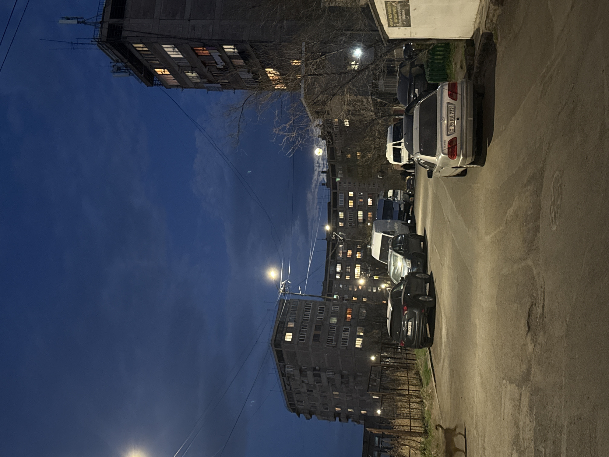 The image depicts a street scene in Yerevan, Armenia at dusk. Several multi-story buildings line the street, some with illuminated windows. Parked cars are visible along the right side of the road. A row of bare trees is present near the buildings. Streetlights are on, illuminating the scene. The sky transitions from a lighter shade at the horizon to a deep blue at the top of the frame.