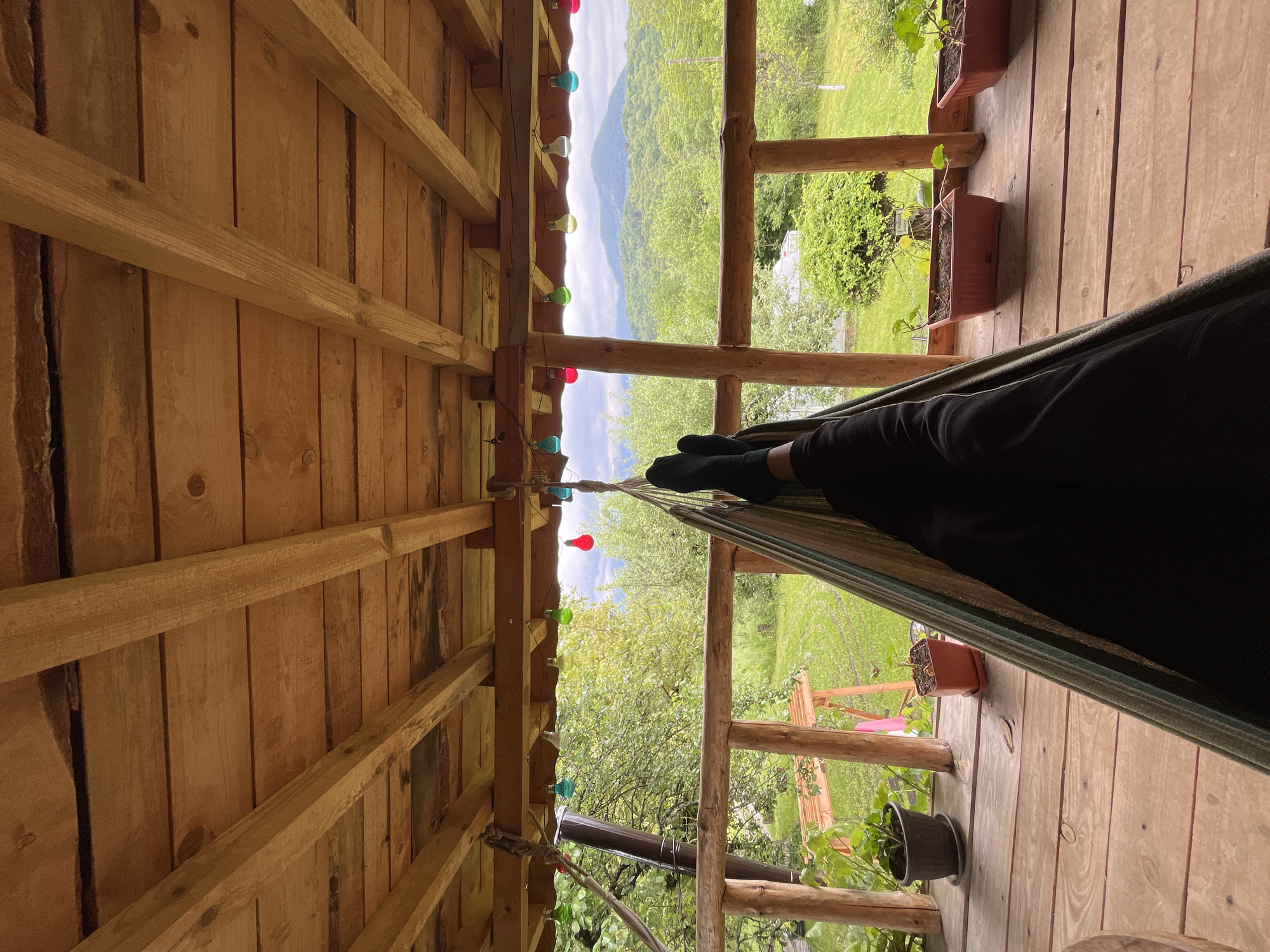 Here's a detailed description of the image:
**Location:** Shopite, Bulgaria.
**Scene:** A person is relaxing in a hammock on a wooden porch or deck of a building. The deck is enclosed by wooden railings. Potted plants are placed along the perimeter. The view from the porch offers a scenic landscape of green hills and a mountain range under a partly cloudy sky.
**Objects:**
* Hammock
* Wooden porch/deck
* Wooden railings
* Potted plants
* Strings of colorful lights hanging from the wooden ceiling beams
* Mountains in the background
**People:**
* A person is partially visible, lying in the hammock, only their legs and feet wearing black socks are visible.
**Environment:**
* The environment suggests a peaceful, natural setting in a rural or semi-rural area. The presence of a wooden structure and surrounding greenery indicate a dwelling or recreational space designed to appreciate the landscape.