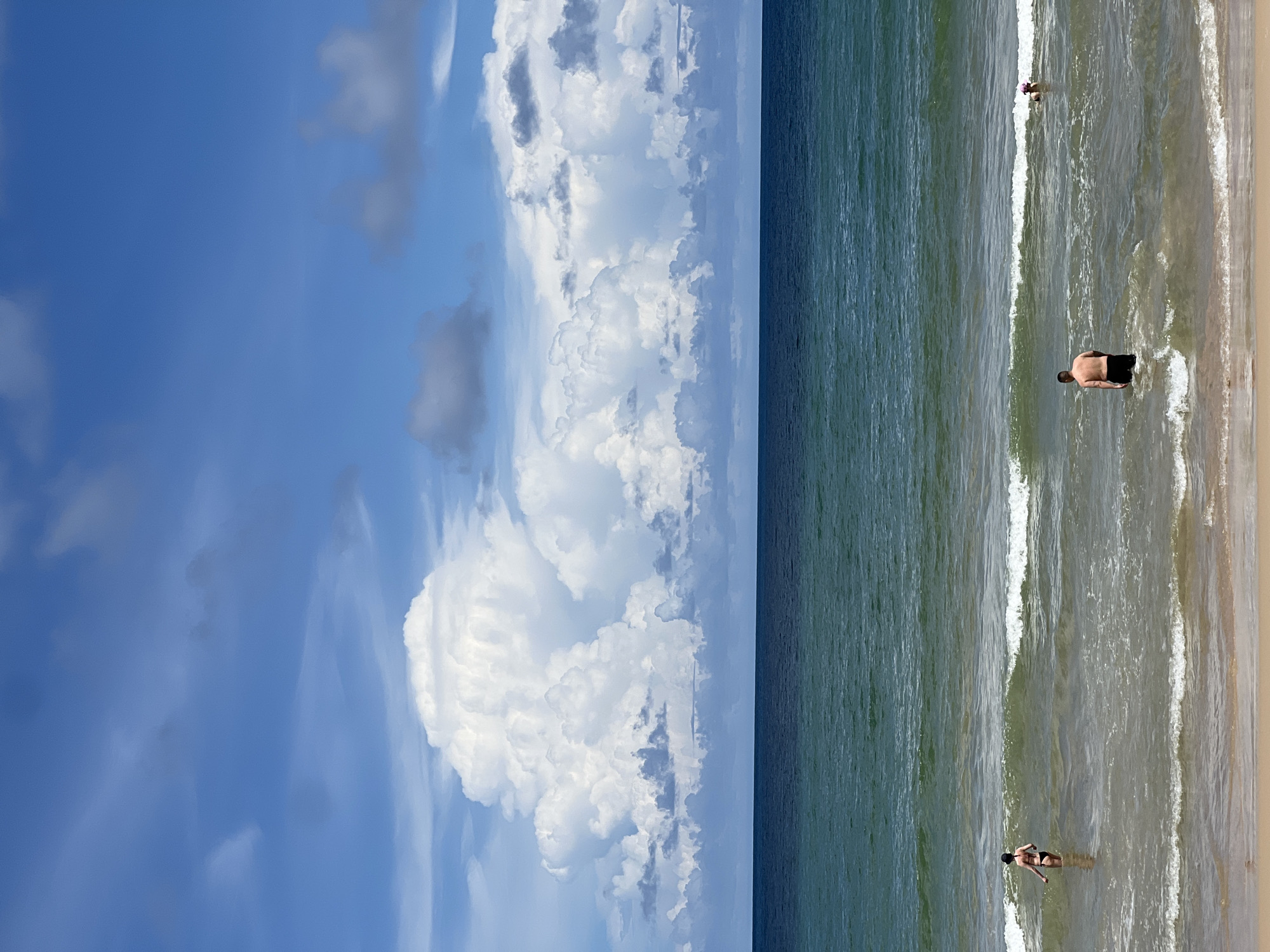 A wide, low-angle shot captures a serene beach scene under a partly cloudy sky. In the foreground, the sandy beach slopes gently into the ocean. Several people are visible wading in the water. A woman in a black bikini stands facing away from the camera, her back and legs submerged. Further out, a man in black swim trunks stands with his back to the viewer, his torso and head visible above the water. A second person, possibly a woman with a pink cap, is also in the water further to the right. The ocean itself is a deep blue, with lines of white surf breaking as they approach the shore. The horizon separates the water from the sky, which is dominated by dramatic, fluffy white cumulus clouds against a bright blue backdrop. The overall impression is one of a peaceful, sunny day at the beach.