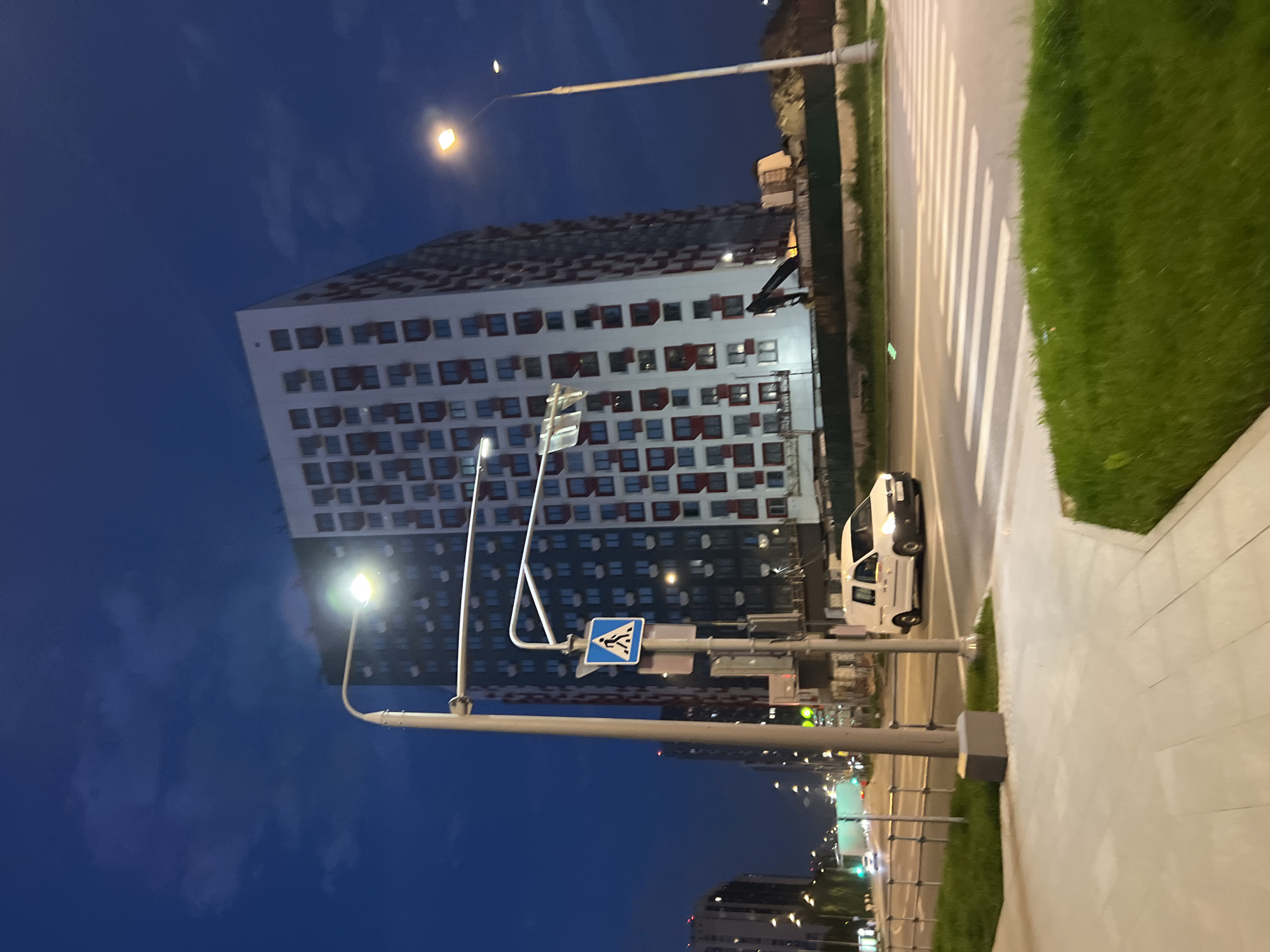 The image shows a nighttime street scene in Vnukovo, Russia. A tall, white apartment building with red window frames dominates the background. A streetlamp illuminates the building's side. A crosswalk with white stripes runs across the road. A white van is driving on the road next to the pedestrian crossing. A pedestrian crossing sign is visible on a pole near the crosswalk. A sidewalk runs along the right side of the image, lined with green grass. Further down the street, city lights are visible. The sky is dark blue with some clouds.