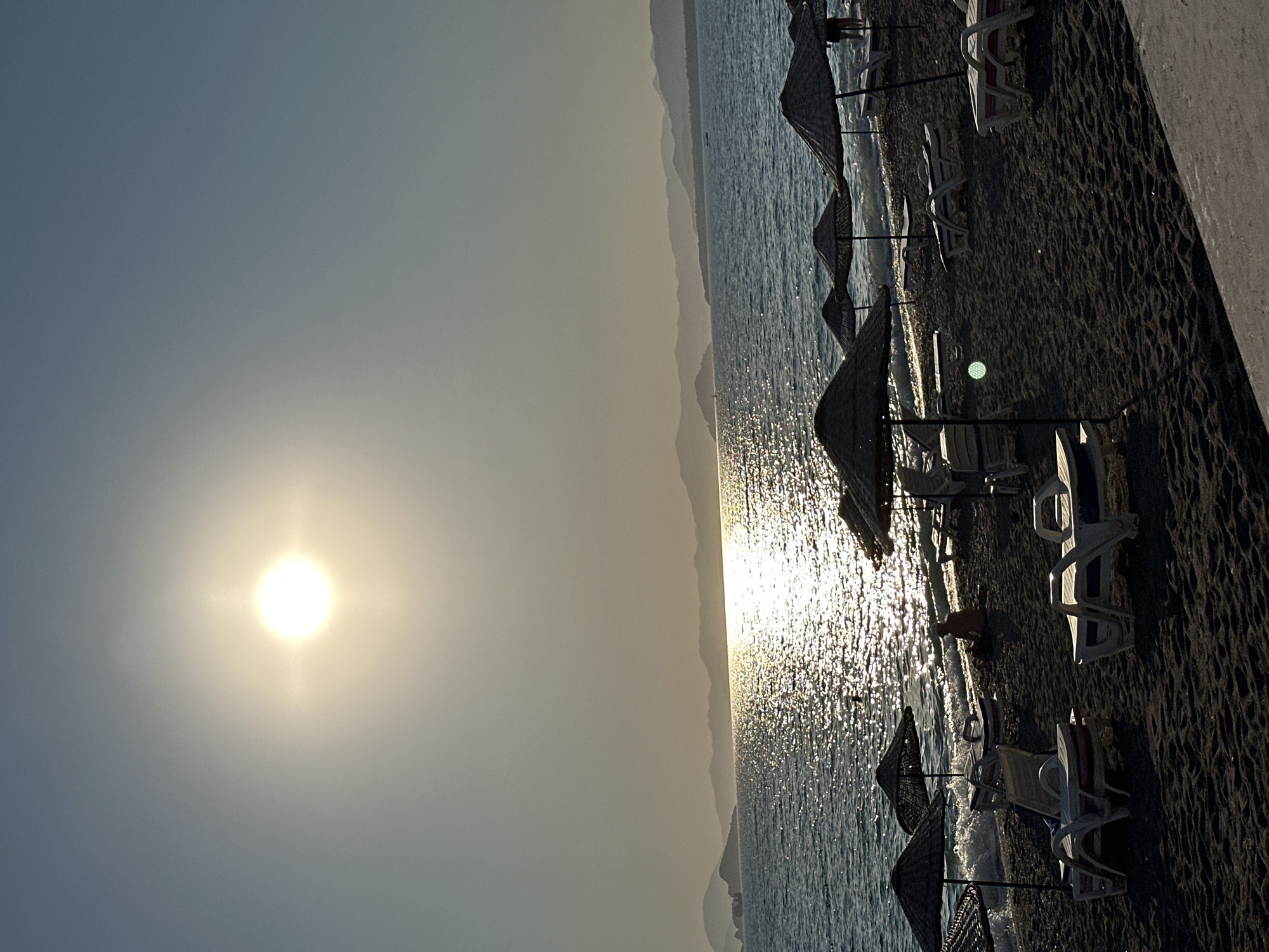 The image depicts a serene beach scene in Fethiye, Turkey, likely during sunset or sunrise. The sun, a bright orb in the sky, casts a glittering path across the tranquil sea. Gentle waves lap at the shore, where rows of empty lounge chairs and straw umbrellas await beachgoers. The sand is dark, suggesting a volcanic or mineral-rich composition. In the background, a chain of hazy mountains forms a picturesque silhouette against the horizon. A lone figure sits on the beach, a small boat sails in the distance, and the overall atmosphere is one of peaceful repose.