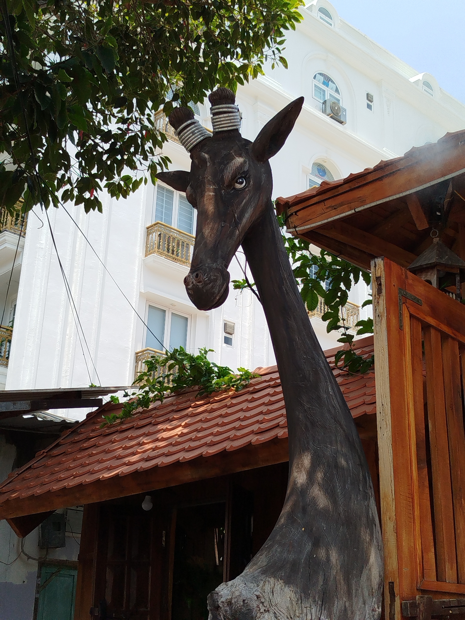 The image depicts a wooden giraffe sculpture in Ho Chi Minh City, Vietnam, standing near a building with a tiled roof and an adjacent wooden structure. The sculpture is dark in color, with striped ornamentation around its horns. It is positioned near foliage, with a white building featuring ornate balconies visible in the background. A wooden birdhouse hangs from the roof of the adjacent structure.