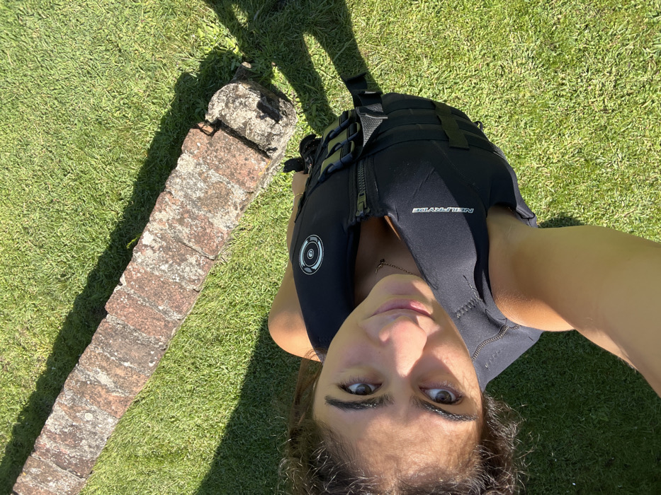 A female subject, appearing to be a young adult, is depicted outdoors in Coruche, Portugal. She is wearing a black NeilPryde brand flotation vest or impact vest, featuring a front zipper, adjustable straps with buckles on the side, and a circular emblem on the upper chest. Her dark, curly hair frames her face, and her brown eyes are directed towards the camera in a self-portrait. The background consists of vibrant green grass and a low, linear structure made of weathered reddish-brown bricks, partially covered in light-colored lichen or moss. Bright sunlight illuminates the scene, casting distinct shadows on the grass from both the subject and the brick structure.