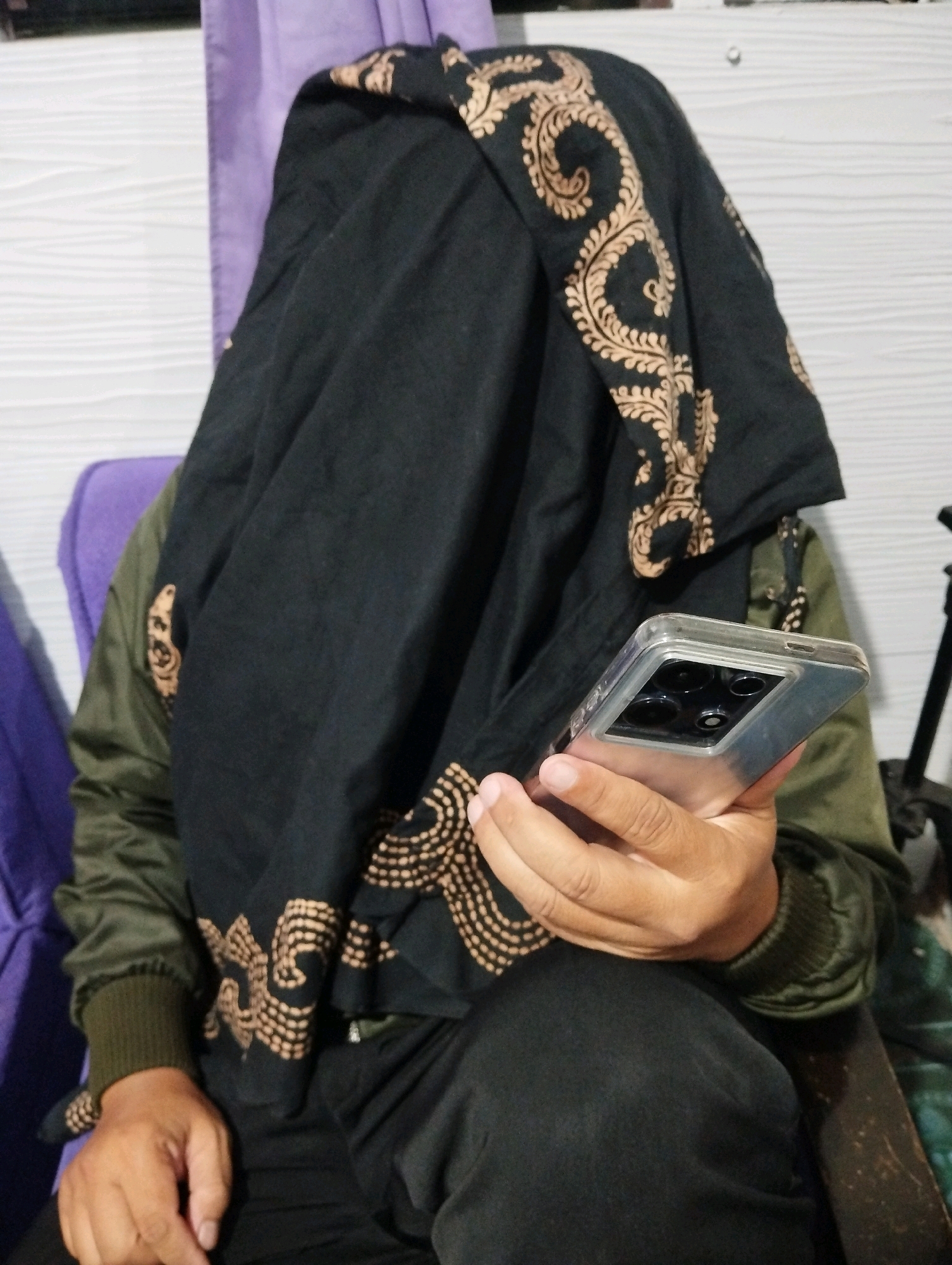 A person is sitting on a purple chair, holding a smartphone. The person is wearing a green jacket and a black hijab with a golden pattern. The background is a white wall and a purple curtain. The image is taken indoors in the city of Salatiga, Indonesia.