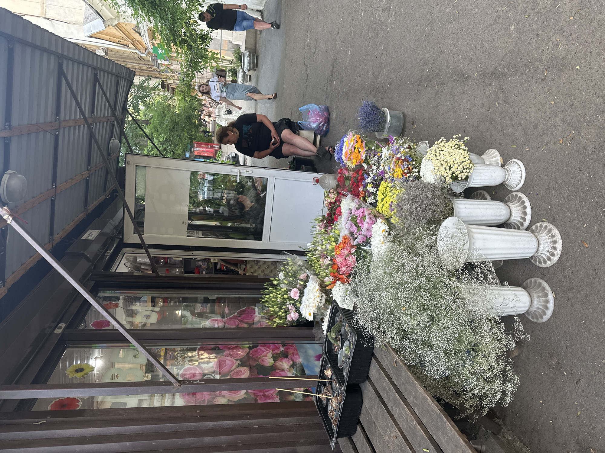 A street-level view captures a flower stall in Odesa, Ukraine, under a cloudy or overcast sky. The scene is oriented vertically, with the top of the image showing buildings and trees, and the bottom revealing the sidewalk and a flower stall.
In the foreground, a woman with dark hair is seated on a bench in front of a flower shop. She is wearing a black t-shirt and black shorts, and appears to be looking down at something in her hands. She is surrounded by a vibrant display of flowers. Bunches of colorful flowers, including yellow, orange, pink, and white varieties, are arranged in front of the stall. Some flowers are in vases, while others are piled high. Small potted plants, including cacti, are also visible on the bench. White ornamental pillars are placed amongst the flowers, adding a decorative element. The flower shop itself has large glass windows, revealing more floral displays inside.
Further back, two other women are standing on the sidewalk. One is wearing a white t-shirt and a light-colored skirt, and the other is in a black t-shirt and blue shorts. A man in a black t-shirt and blue shorts is also visible standing near the buildings in the background. The people appear to be casually interacting, suggesting a normal day at the market.
The setting appears to be an urban street with buildings lining one side and the sidewalk and flower stall on the other. The presence of trees suggests a temperate climate. The overall atmosphere seems relaxed and ordinary, with the focus on the bustling flower market. No visible text is discernable in the image.