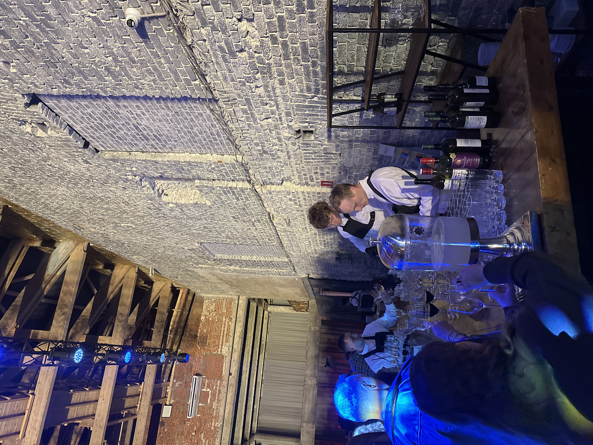 The image shows a dimly lit, indoor setting, possibly a bar or event space with exposed brick walls and wooden beams. The camera is angled vertically, making it appear as if the scene is upside down. There are several people visible, mostly from the shoulders up, and they appear to be serving drinks. Two individuals in white shirts and black vests are at a bar, interacting with glassware and a large silver beverage dispenser. One person is wearing a blue illuminated jacket or shirt, with their head facing away from the camera. Another person in the foreground has bright blue hair and is also facing away.
On a wooden shelf behind the bar, several bottles of wine are neatly arranged. The lighting is dramatic, with blue spotlights illuminating parts of the scene, particularly the bar area and the people. There is a security camera visible on the ceiling in one of the cropped images. The overall atmosphere appears to be social and possibly celebratory, given the bar setting and the activity of serving drinks. The time of day is difficult to determine due to the artificial lighting, but the ambiance suggests it could be evening or night. There is no visible weather information, and the location is not specified, but the industrial-chic decor might suggest an urban venue.