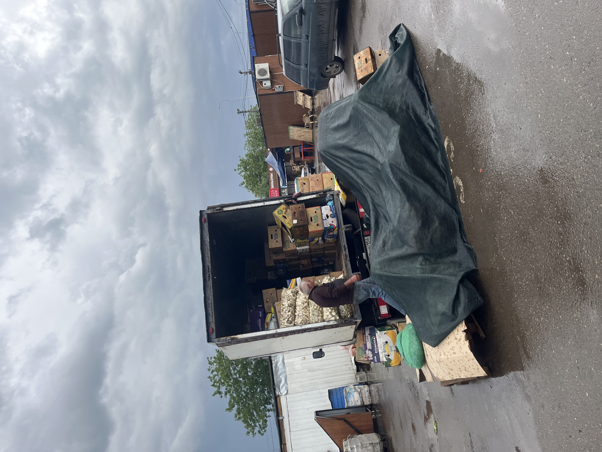 A medium shot captures a loading truck with its back open, filled with boxes and sacks of what appear to be vegetables. A man is standing inside, seemingly organizing the load. The truck is parked beside a low white building. To the right, an object is covered with a dark green tarp. The ground appears wet, suggesting recent rain. In the background, there are other buildings, and a car is parked. The sky is overcast, giving the scene a dim light. A store in the city of Krasnodar, Russia, can be seen in the background.