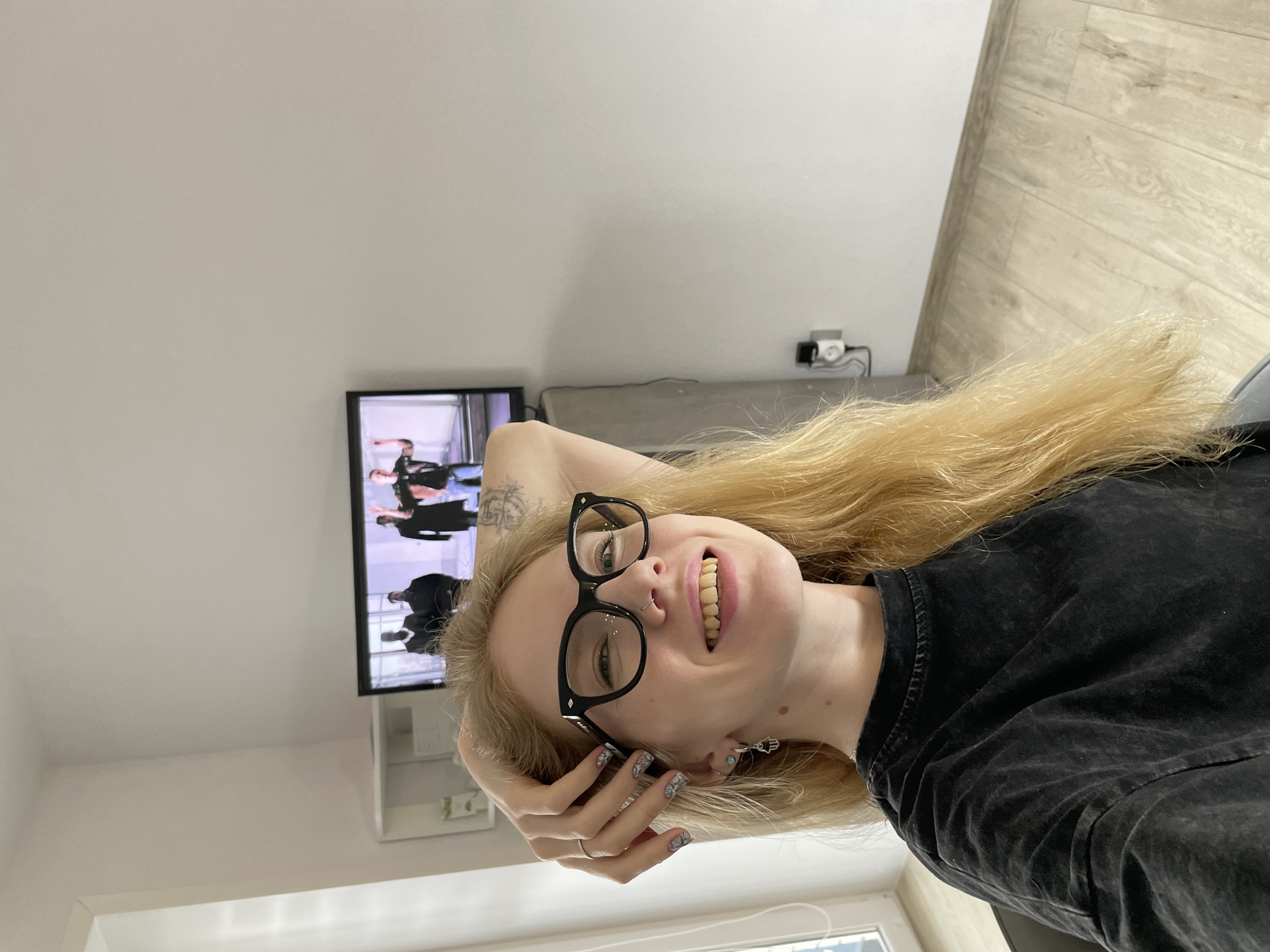 The image shows a smiling young woman taking a selfie indoors, rotated 90 degrees clockwise. She has long, straight blonde hair and is wearing black-framed glasses, a dark t-shirt, and a nose ring. Her left hand is raised to her head, with a silver ring on her ring finger and patterned nail polish visible. She wears a small stud earring in one ear and a silver Hamsa hand charm in the other. A tattoo is visible on her right arm.
The background consists of plain, light-colored walls and a light wooden floor with planks laid diagonally. A flat-screen television is mounted on the wall, displaying what appears to be a scene with several people. Below the TV, there is a small white floating shelf or cabinet. An electrical outlet with a white plug and cables is visible on the wall to the right of the TV. The overall setting appears to be a room, possibly in a home, in Izhevsk, Russia. No discernible text is visible in the image. The woman appears happy and relaxed.