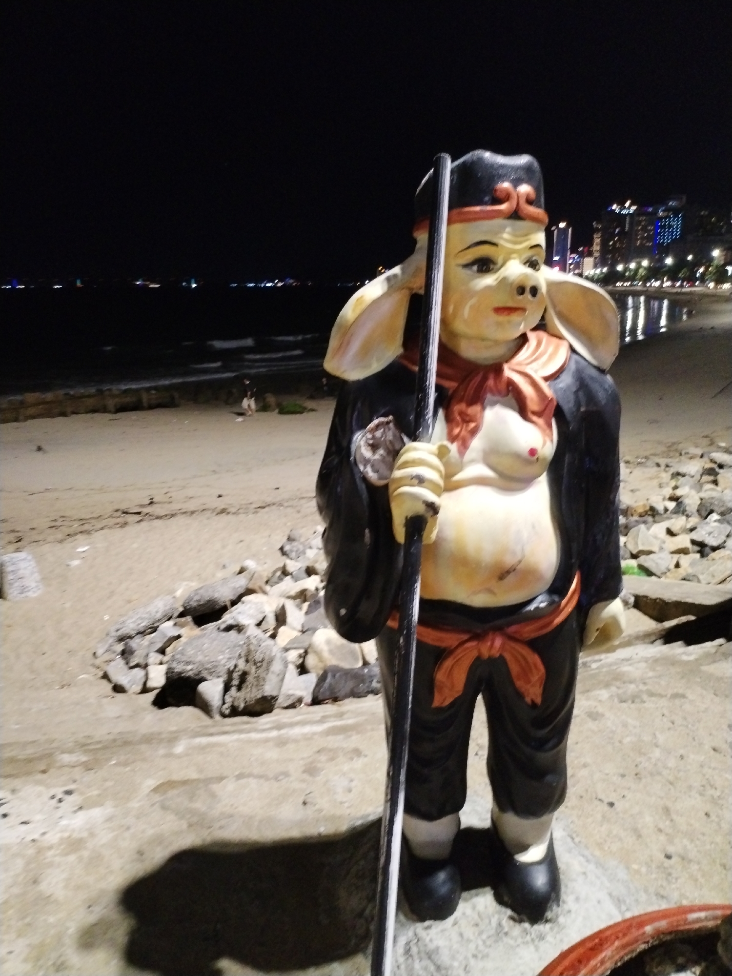 A statue resembling Zhu Bajie, also known as Pigsy from the Journey to the West, is located on a beach in Nha Trang, Vietnam, at night. The statue is standing on a concrete base near a rocky area on the sand. In the background, there is a beach, the ocean, and city lights. There are a couple of people on the beach in the mid-ground. The statue is wearing a black outfit with an orange sash and is holding a staff.