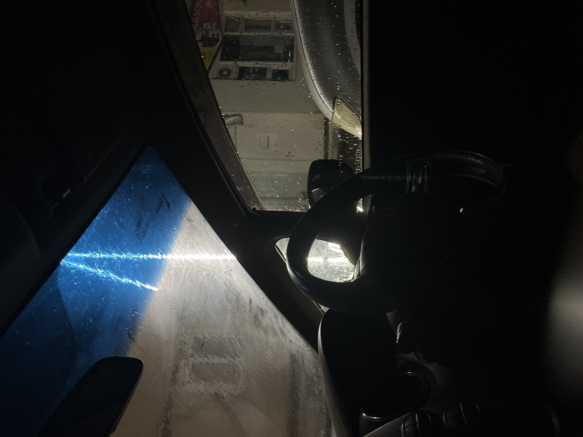 Inside view of a car at night, likely at a car wash in Poti, Georgia. The windshield is wet with water droplets, and streaks of blue and white light are visible on it, possibly from the car wash equipment. The steering wheel is in the foreground, silhouetted against the light. Outside the car, through the wet side window, a building with windows and a silver car can be seen. The scene is dark and atmospheric, suggesting a rainy or humid night.