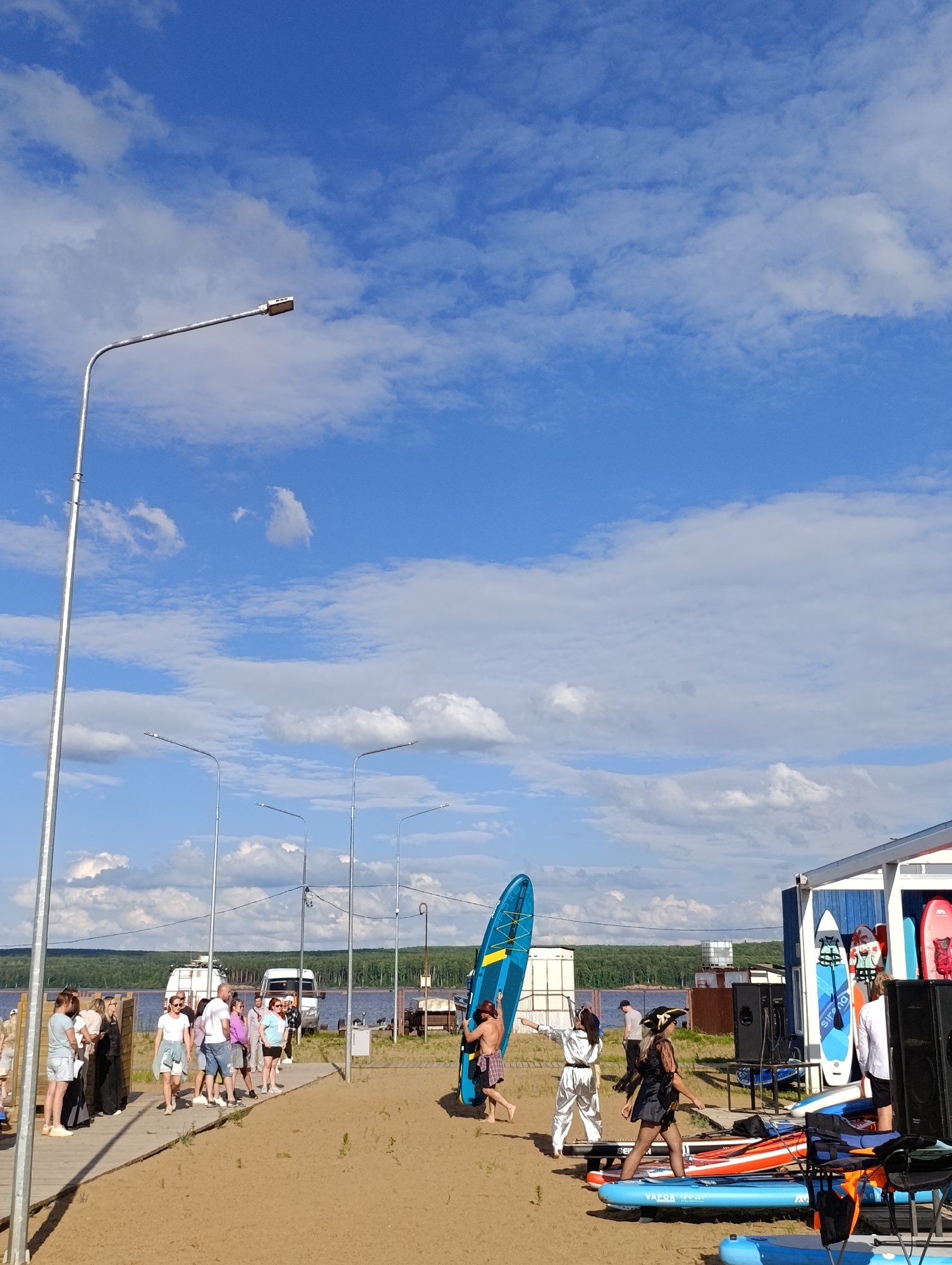 This media file captures an outdoor event on a sandy beach with a body of water in the background, likely a lake or river. The sky is bright blue with scattered white clouds, suggesting a pleasant, possibly sunny day. Several streetlights are visible, implying the area is equipped for public use, and the presence of a building on the right further indicates a developed recreational spot.
The scene is populated by a group of people, some of whom appear to be participating in or observing an activity involving paddleboards. On the right side of the image, several paddleboards are lined up, some leaning against a blue and white structure. A person is holding a large blue paddleboard upright, seemingly posing or preparing it for use. Other individuals are gathered on a wooden walkway, some watching the scene unfold, while others are in motion.
There are hints of a costume or themed event, with at least two people dressed in distinct outfits: one in a silver, shiny outfit and another in what appears to be a pirate costume, complete with a hat. This suggests a playful or festive atmosphere. The people are casually dressed, consistent with a warm weather outdoor event.
The overall mood seems relaxed and social, with people interacting and enjoying the outdoor setting. The presence of vans in the background and the general setup suggest a public gathering or organized activity at a waterfront location. The time of day appears to be afternoon, given the bright sunlight and the angle of shadows. The location context is Chaikovsky, Russia.