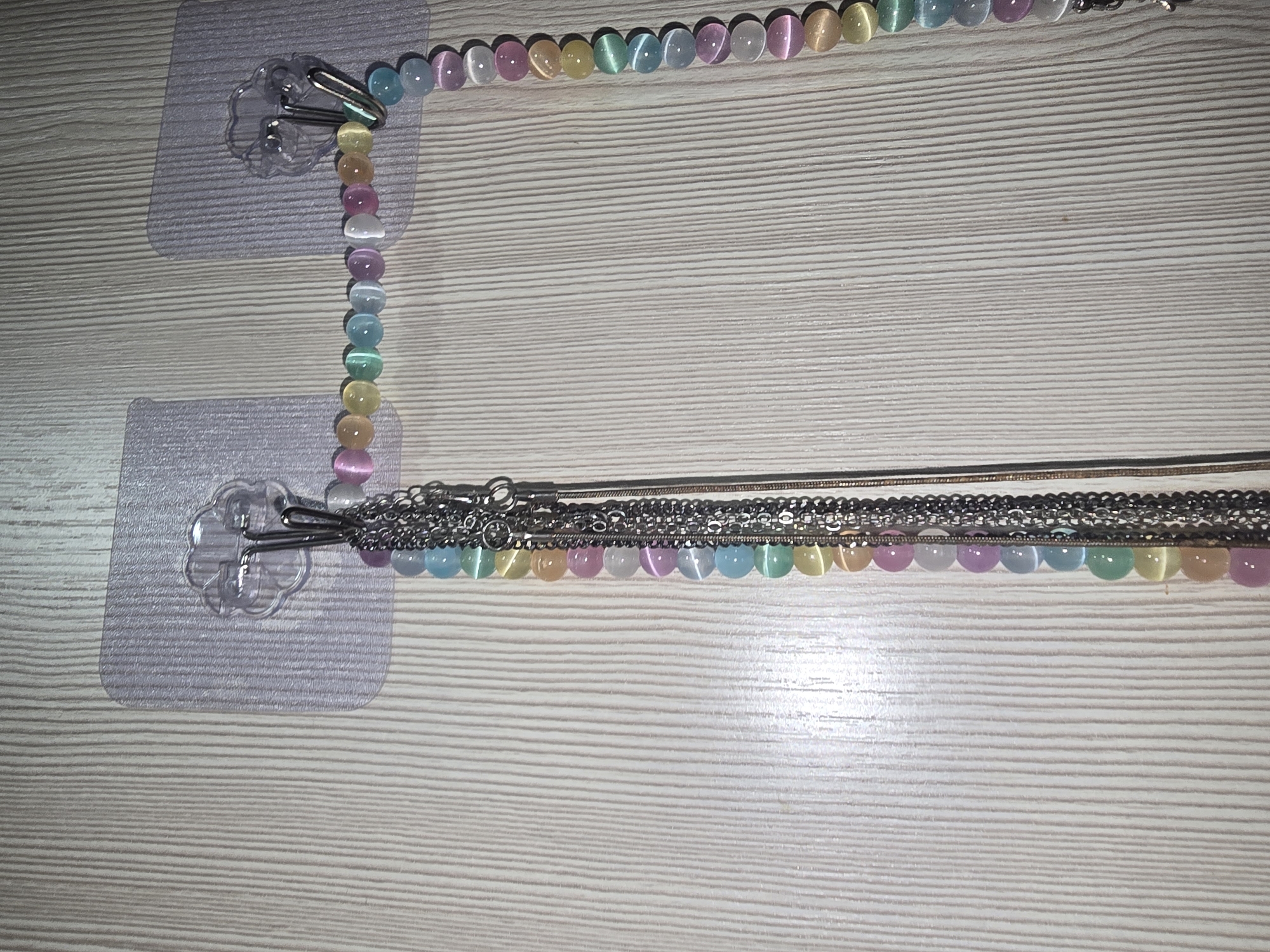 The image shows two clear plastic adhesive hooks mounted on a light-colored wooden wall with vertical grain. Each hook has a floral-shaped edge. Hanging from the left hook are several delicate silver chains. Hanging from the right hook is a multi-colored beaded necklace composed of round, iridescent beads in shades of pink, orange, yellow, green, and blue. The necklace is draped in a U-shape, with both ends of the necklace attached to the right hook. The lighting appears to be from above, casting soft shadows. There are no people or animals present. The scene depicts a simple organization of jewelry. The image is not specific to Ulan-Ude, Russia, and provides no contextual information about its location.