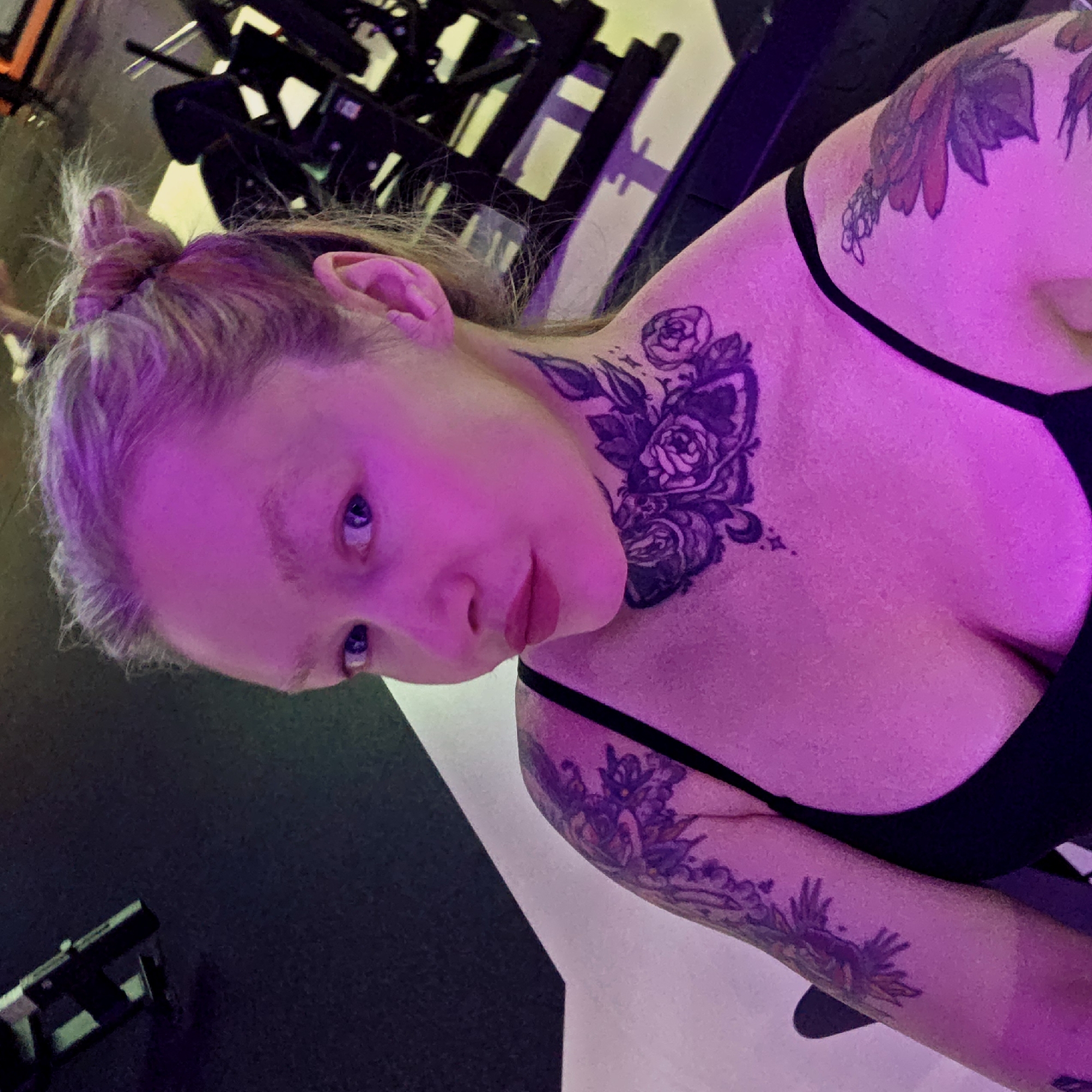 An individual with light-colored hair, tied up in a bun, is captured in a close-up selfie within an indoor setting in Saint Petersburg, Russia. The person, likely female, wears a black top or sports bra and has multiple tattoos visible on their neck, shoulder, and forearm, featuring intricate floral and organic designs.
The scene is dominated by strong artificial purple and pink lighting, which casts a vivid magenta hue across the person's skin and much of the visible environment. Their eyes, a light color, reflect the surrounding light. In the blurred background, dark structures resembling gym equipment or weight racks can be discerned, suggesting a fitness studio or similar facility.
The person looks directly at the camera with a neutral to slightly contemplative expression. The image is taken from a tilted angle, giving it a dynamic composition. There is no visible text within the media file.