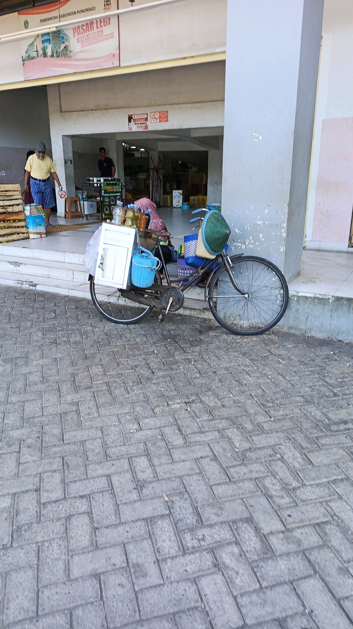 In front of a market stall in Nologaten, Indonesia, a bicycle with a white cargo box is parked on a brick-paved sidewalk. The bicycle is laden with goods, including several bottles of liquid, a blue bucket, and a green conical hat perched on the handlebars. A person wearing a pink hijab is seated behind the bicycle, and a man in a yellow shirt and checkered shorts stands to the left of the frame, holding a small item. Behind them, the market features stalls with produce and a sign indicating "PASAR LEGI." The environment suggests a bustling outdoor market scene.