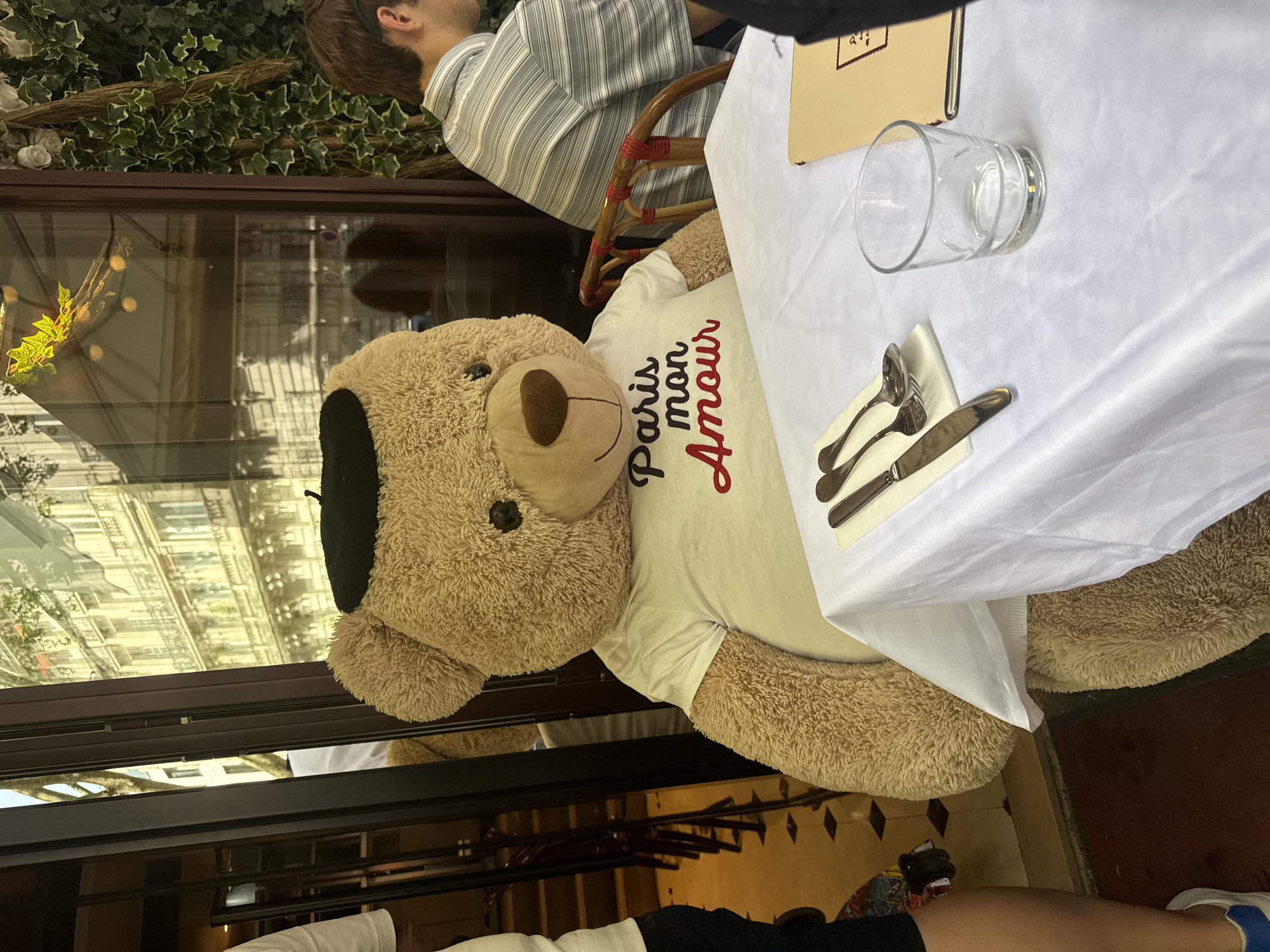 A sideways indoor/outdoor shot shows a teddy bear wearing a beret and a T-shirt that says "Paris Mon Amour" sitting at a table with silverware and a glass in what appears to be an outdoor cafe setting. The cafe is partially enclosed by a window, and a building is visible through the window's reflection, with a person with short hair wearing a striped shirt sitting behind the bear. The floor tiles and table style suggest that the photo was taken in a Parisian cafe.