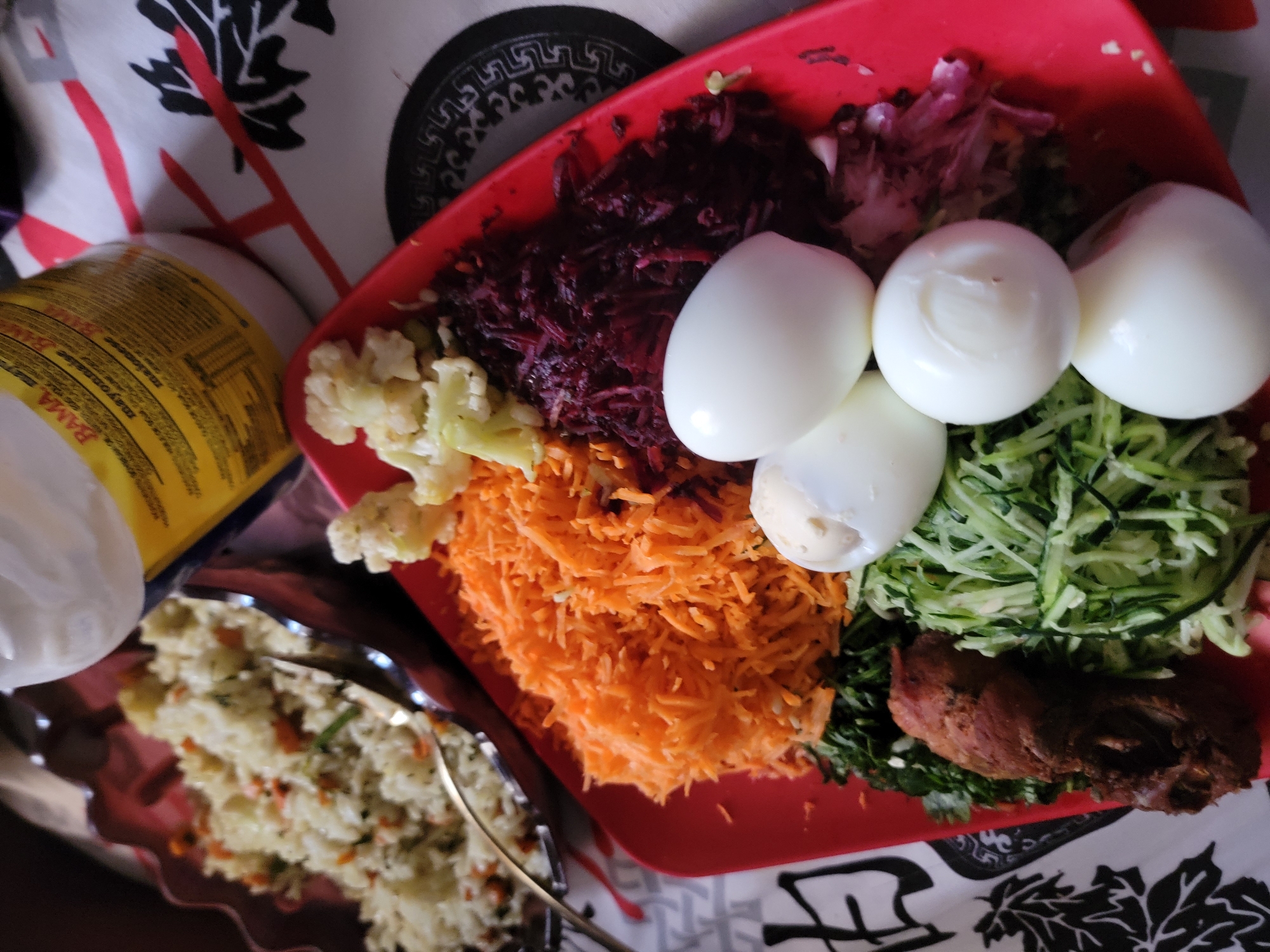 A top-down view shows a red plate containing several types of food arranged in sections. These include shredded beets, hard-boiled eggs, shredded carrots, and shredded zucchini. A piece of cooked meat sits in a lower corner near some green leaves. A second silver plate contains a rice-based dish. A container of mayonnaise sits to the left. The plate and dishes are on a patterned tablecloth with black leaves on a white background. The location of the image is at Jos, Nigeria.