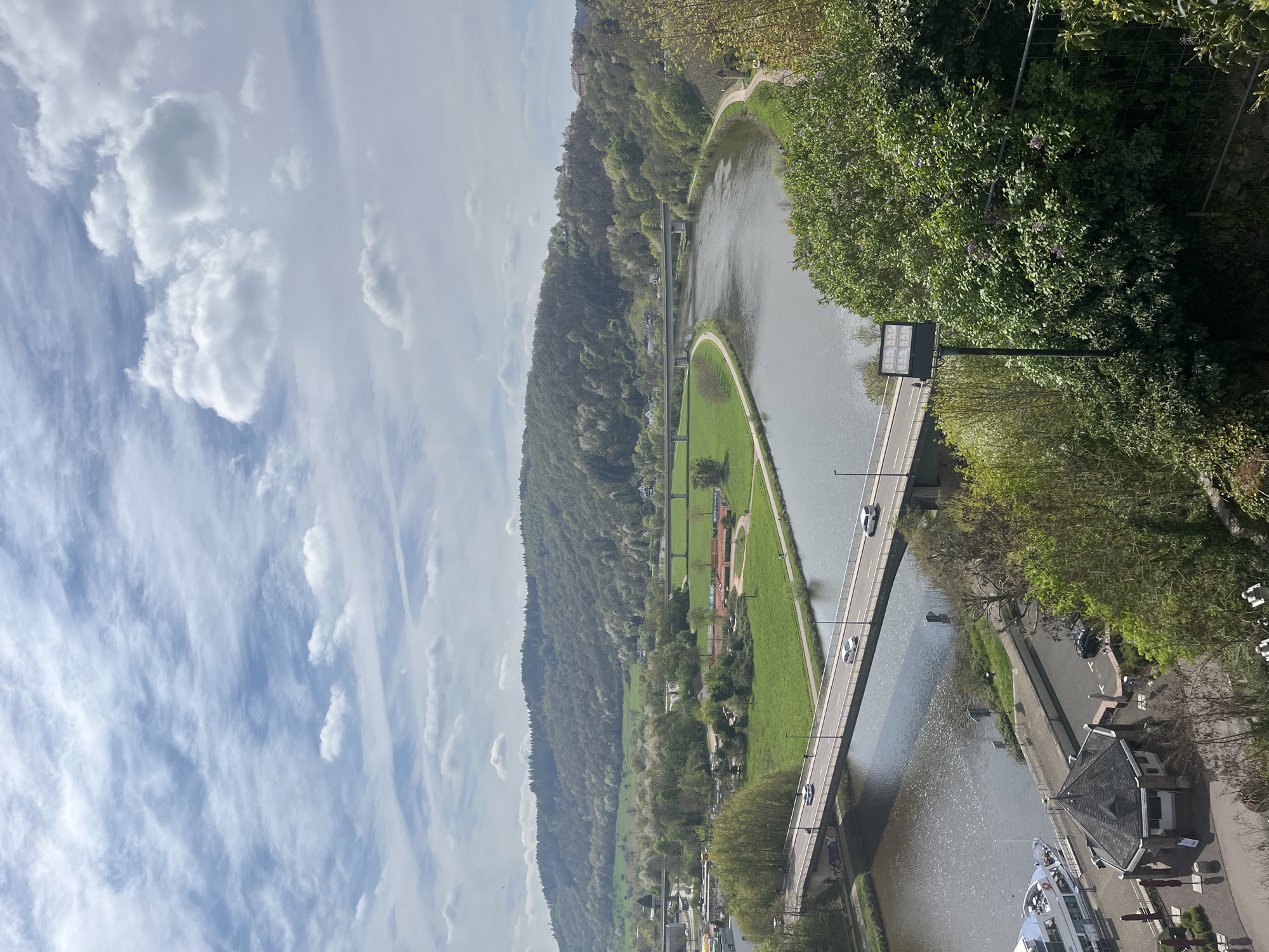 The image is a vertical aerial view of Saarburg, Germany, featuring a river with a bridge spanning it. The landscape is marked by a tree-covered hill on one side and buildings on the other. Cars are visible on the bridge. The sky is partly cloudy. There's a narrow strip of green land between the river and the hill. A white boat is visible near the buildings.