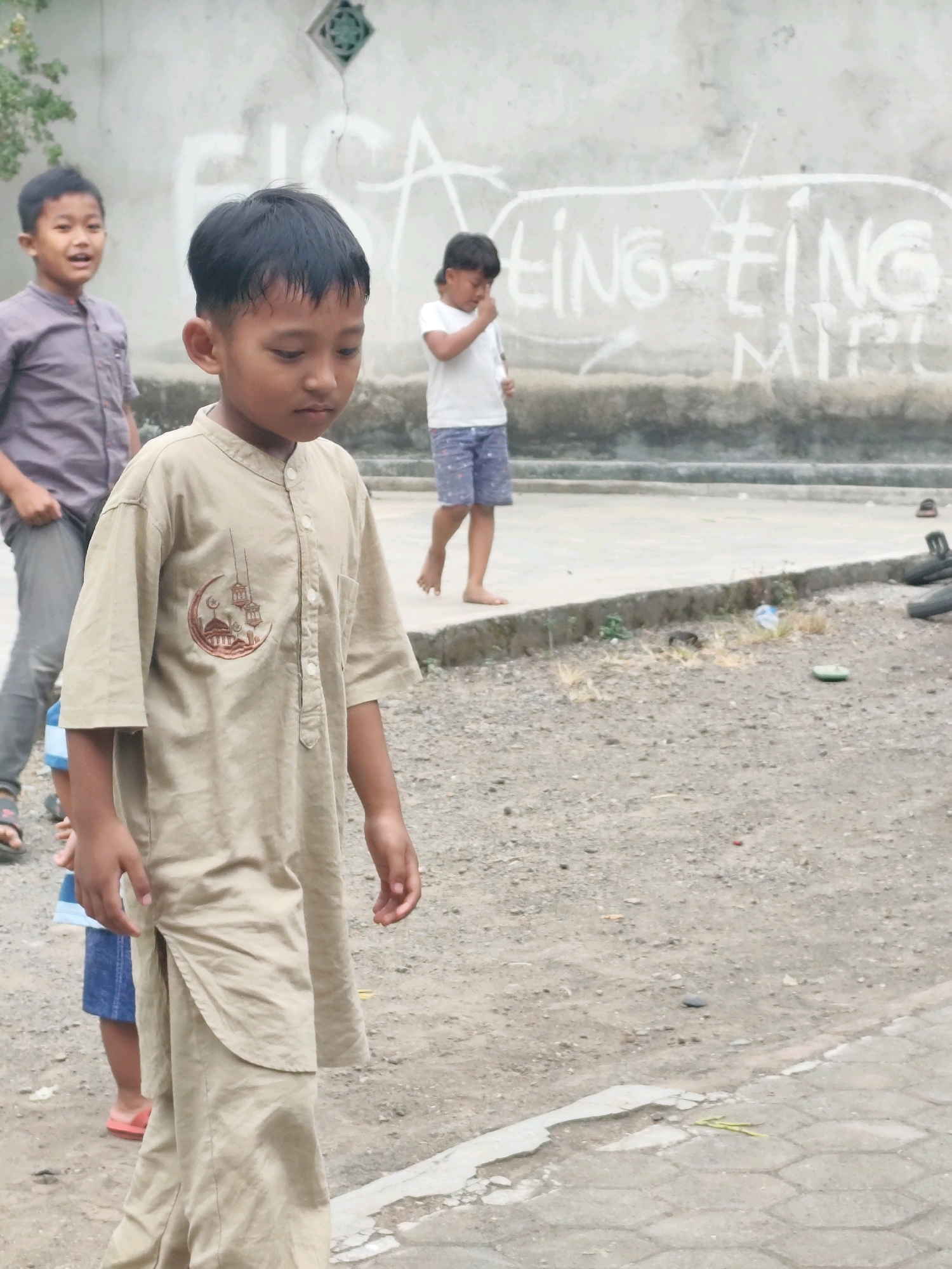 In Tegalsari, Indonesia, a group of children are standing on a dirt and concrete surface in front of a concrete wall. The child in the foreground, wearing a tan tunic with a crescent moon and mosque design, is looking down. To the left, another boy in a grey collared shirt and grey pants is looking towards the viewer with his mouth slightly open. Further back, a third child in a white t-shirt and patterned shorts is looking down and touching their face. The wall behind them has graffiti on it. The ground is littered with small stones and debris.
