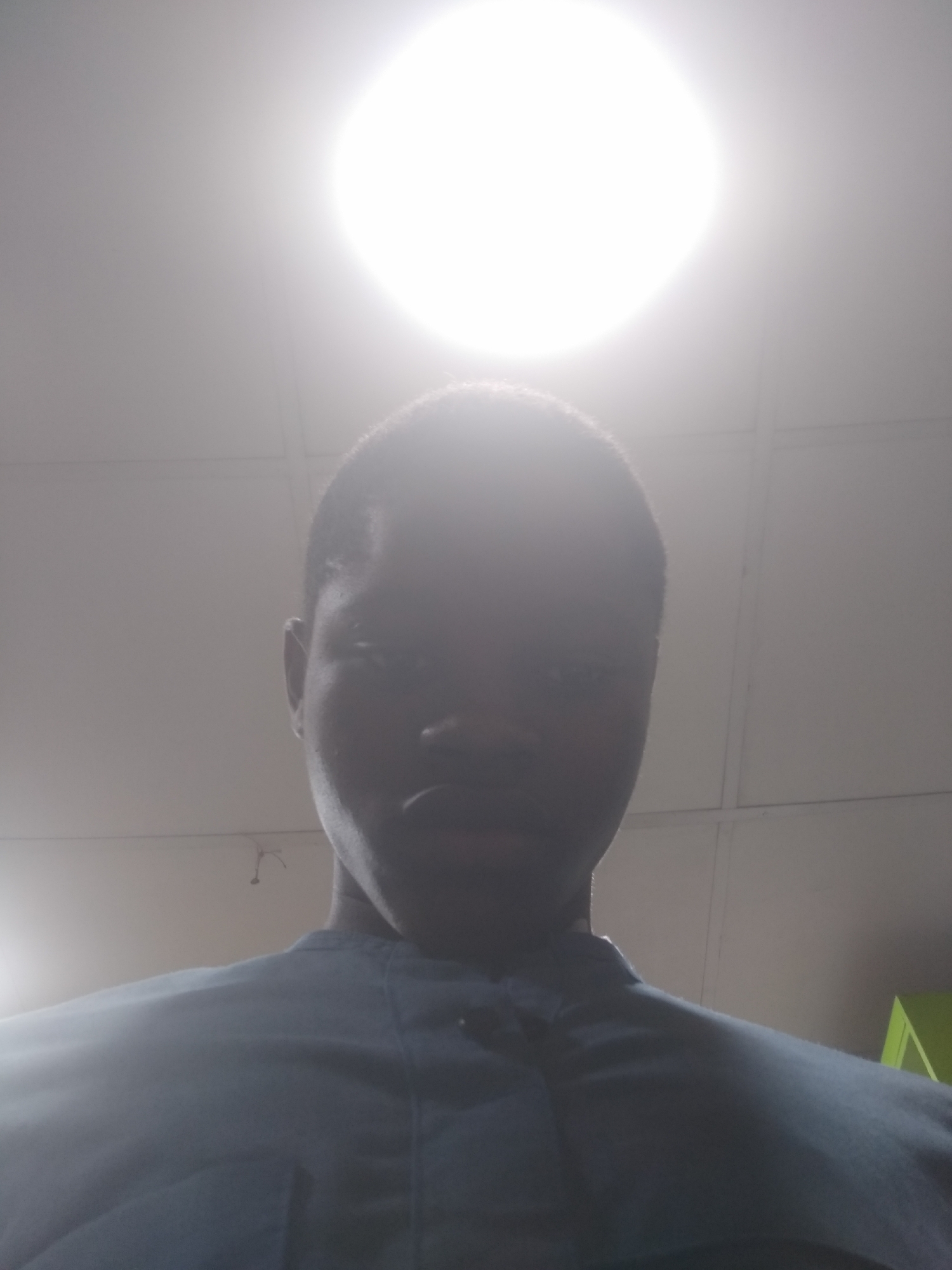 A person is positioned underneath a bright overhead light fixture. The person is facing directly toward the camera with their head tilted slightly downward. The person has a dark complexion and is wearing a dark colored shirt. The background appears to be an indoor space with a light-colored ceiling. The location is not verifiable from this image.