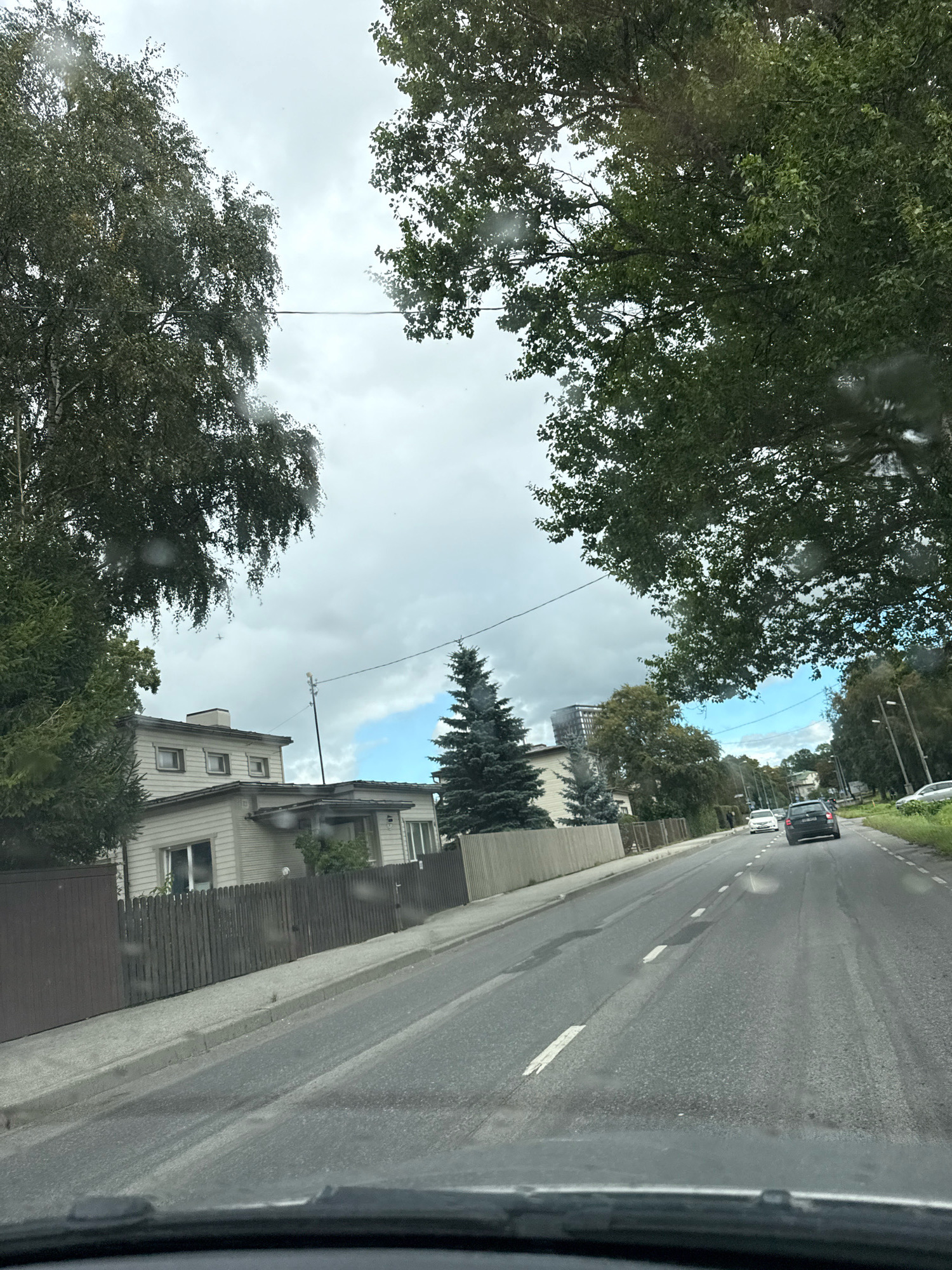 The image shows a view from a vehicle driving down a road in Tallinn, Estonia. The road is paved and has lane markings. On the left side of the road, there are houses with fences and trees. On the right side of the road, there are more trees and some streetlights. There are cars on the road, one of which is a dark-colored SUV. The sky is cloudy, but there are patches of blue sky visible. The overall impression is of a residential area on an overcast day.