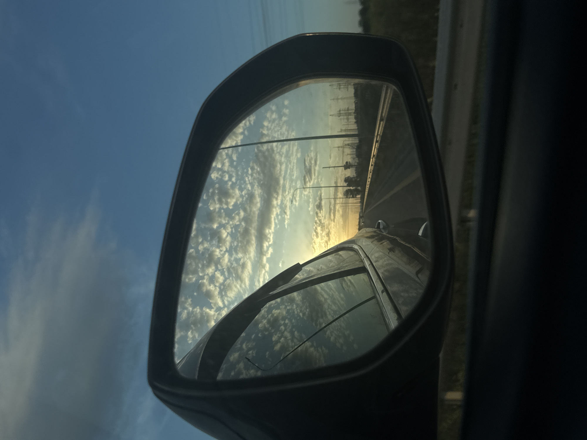 The image is a view through a car's side mirror. The mirror reflects a scene of a highway with power lines and lamp posts along the side. The sky is visible above the highway, with a mix of blue and fluffy clouds. The setting appears to be at sunset or sunrise due to the warm light. The car's body is visible within the mirror's reflection as well. The location of Starotitarovskaya, Russia, is not discernible from the image.