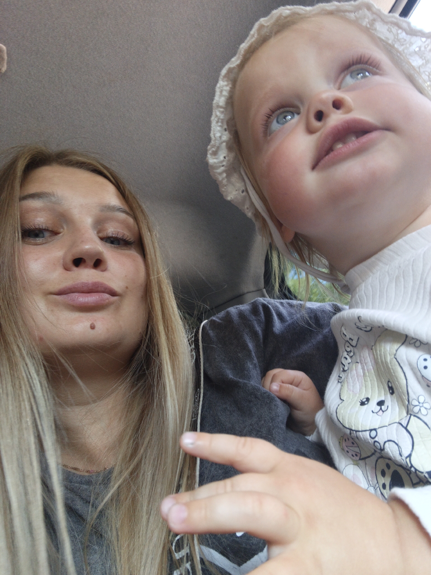The image depicts two individuals inside a vehicle. On the left is a woman with blonde hair, looking down and slightly to the side. Her face is illuminated, and she appears to be taking a selfie. To the right, a young child wearing a white bonnet and a white shirt with a cartoon bear print is looking upwards. The child's face is also visible, with bright blue eyes and rosy cheeks. The background shows the interior of a car, with the ceiling visible at the top. The overall lighting suggests it is daytime, possibly overcast. There is no identifiable information within the image that places it specifically in Zlatoust, Russia.