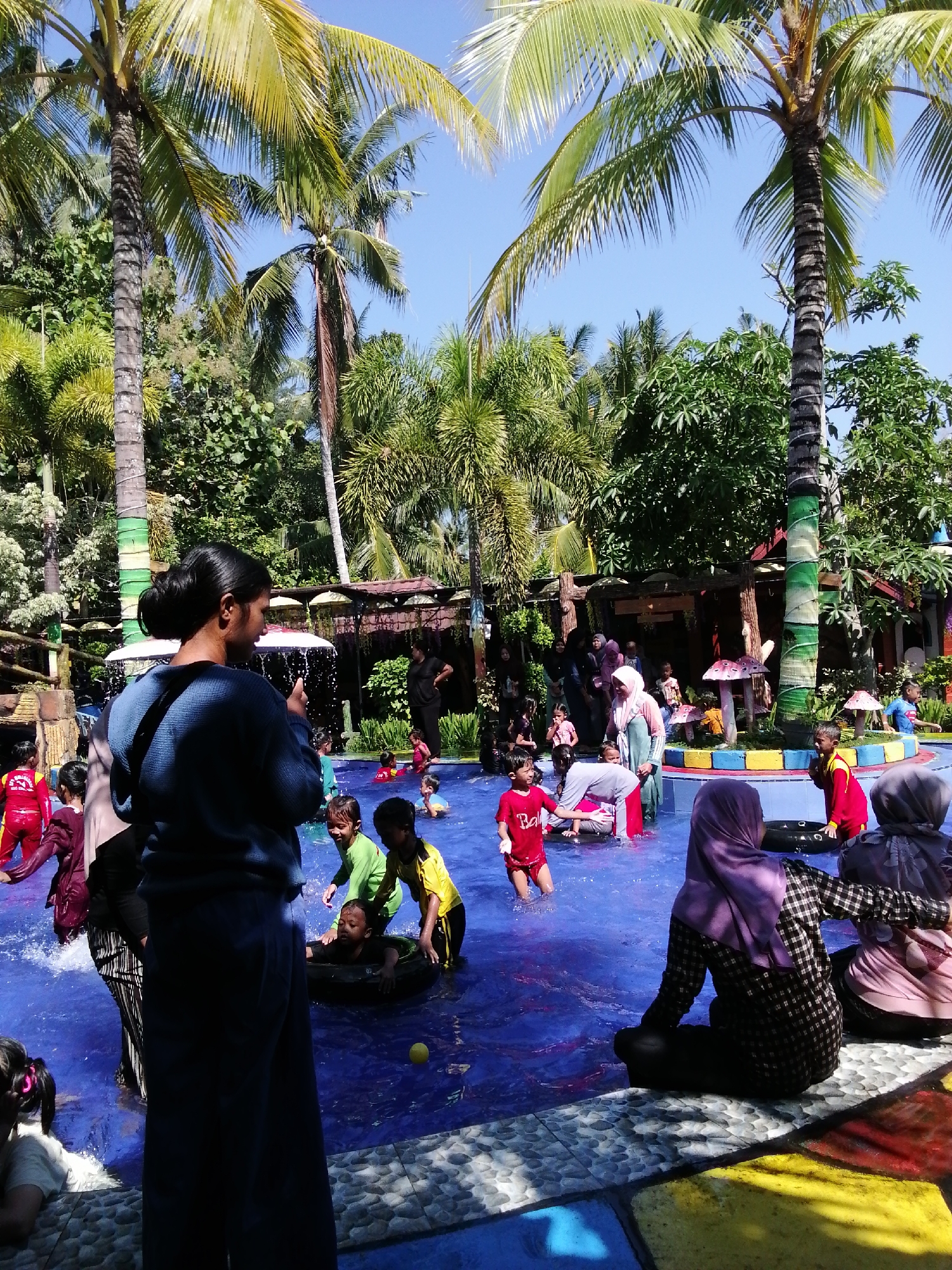 The image depicts a lively outdoor swimming pool scene in what appears to be a water park. The pool has bright blue water and is populated with numerous individuals, predominantly children, engaged in various water activities. Several children are splashing, playing, and swimming, with some using inflatable rings. Adults are also present, supervising and participating.
The environment is tropical, characterized by lush green vegetation and tall palm trees that cast shadows across the scene. In the background, there are structures that suggest amenities like a covered seating area or changing rooms, decorated with colorful, whimsical mushroom-shaped elements. The ground surrounding the pool is a mosaic of colorful tiles in shades of blue, yellow, and red. The sky is clear and blue, indicating a sunny day.
While the image doesn't explicitly state the location as Tegalwangi, Indonesia, the presence of tropical flora, local attire on some individuals (such as headscarves), and the general aesthetic are consistent with such a setting in Indonesia. The overall impression is one of a fun and recreational family outing at a water park.