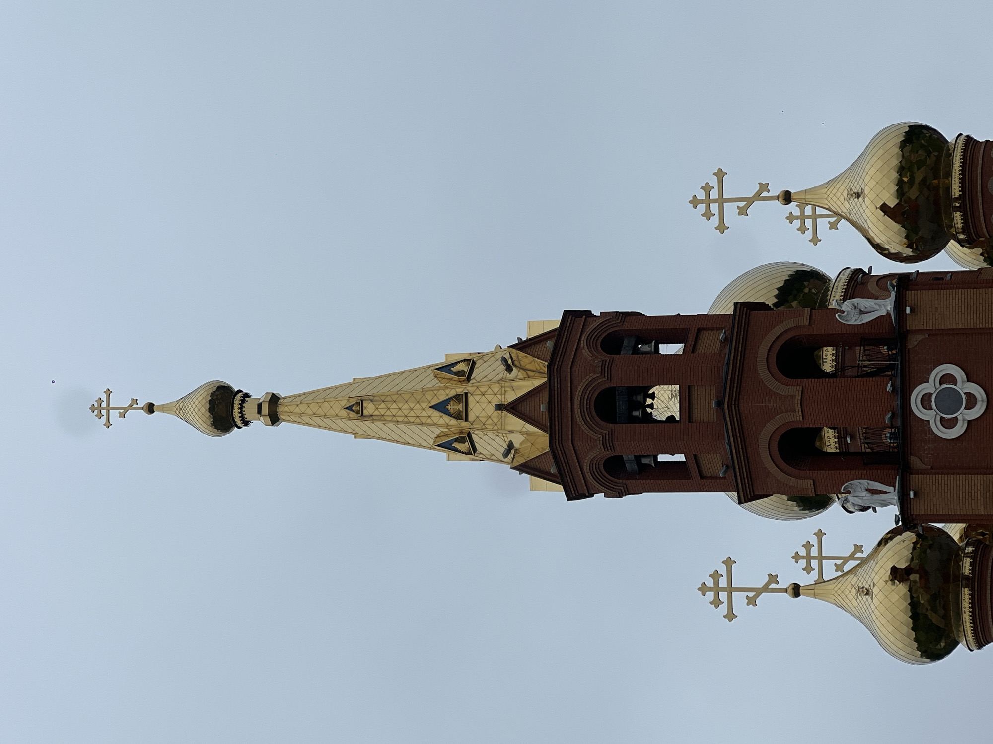 This image showcases a striking Eastern Orthodox church building, prominently featuring its intricate architecture against a light, overcast sky. The central element is a tall, golden-roofed steeple, tapering upwards with a multifaceted design and smaller triangular dormers. At the apex of this spire sits a delicate golden onion dome, surmounted by a distinctive Orthodox cross.
Flanking the main steeple are several larger, more traditional golden onion domes, each crowned with a three-barred Orthodox cross. The main body of the church visible beneath these golden structures is constructed from rich red brick, characterized by decorative arches. These arches likely form part of a bell tower, with some internal structures and bells dimly visible within the openings. On the brickwork, near the base of some domes, several white statues of angels are intricately placed, adding to the church's ornamentation.
Within one of the arched brick openings, a small, blurry inscription in white Cyrillic script can be faintly discerned against a dark background, appearing to include the archaic Russian word "АЗЪ" (Az). The sky is a uniform, pale blue-gray, indicating an overcast day or diffused sunlight. No specific event or activity is taking place; the image is a static architectural portrait. The design, including the onion domes and Orthodox crosses, strongly identifies it as a Russian Orthodox church, consistent with the provided location context of Chaikovsky, Russia.