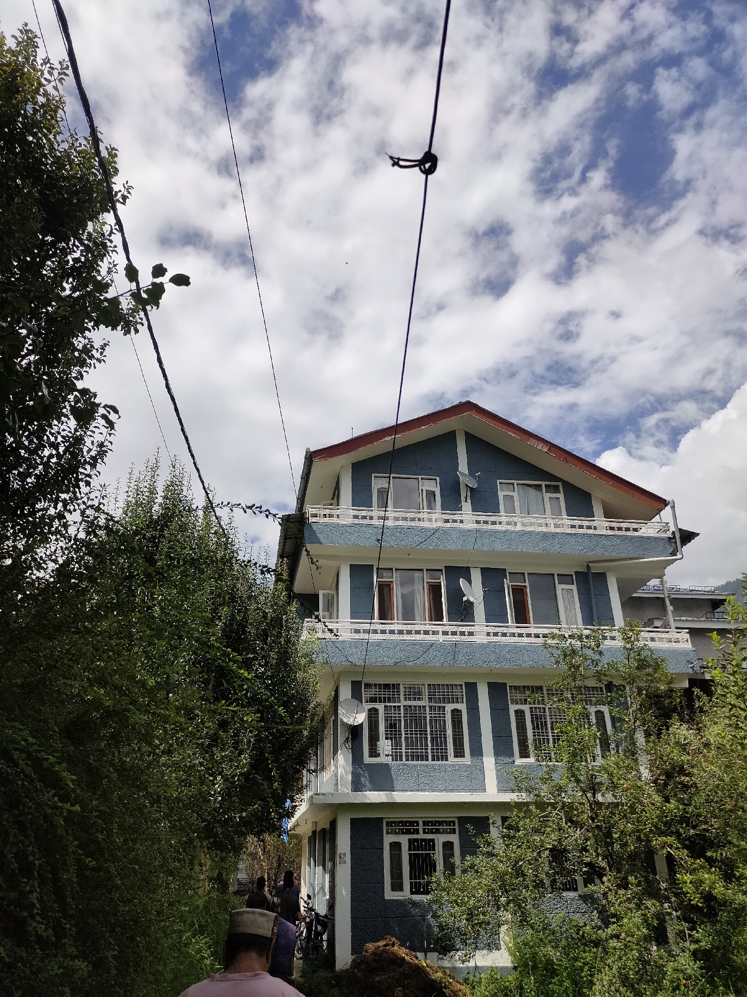 The image depicts a multi-story blue building situated in Manali, India. The building has white trim around windows and balconies, and a red-tiled roof. Several satellite dishes are visible on the exterior walls. In the foreground, lush green trees and foliage line a pathway. A few people are walking away from the camera, with one individual in the foreground wearing a light-colored cap. Overhead, power lines crisscross the frame, and a cloudy blue sky is visible. The scene appears to be an outdoor residential or hotel area, common in tourist destinations like Manali.