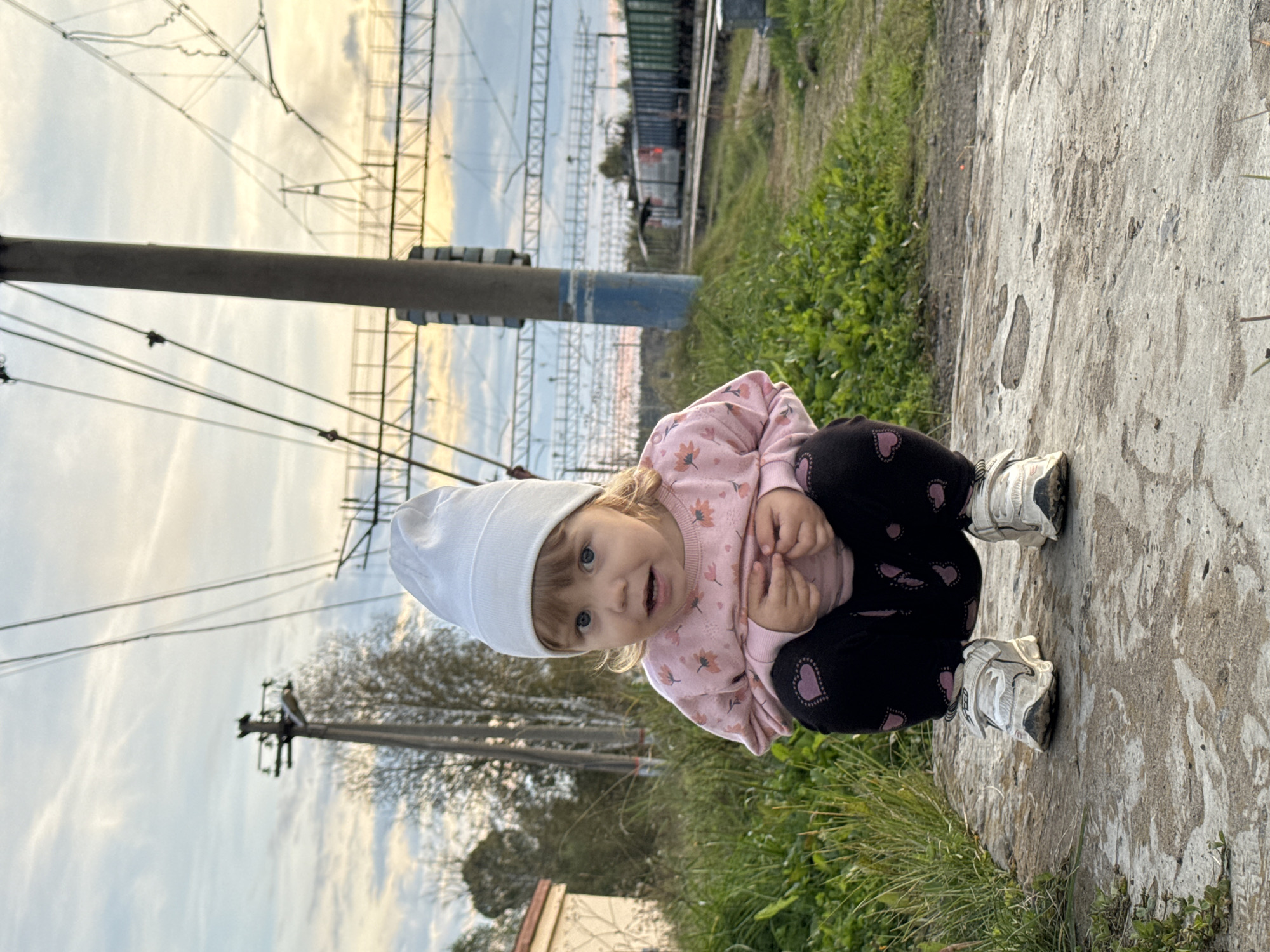A young child, wearing a white beanie, a pink sweatshirt with floral patterns, black pants with heart designs, and white sneakers, crouches on a concrete surface. The child is looking directly at the camera with a curious expression. In the background, there are power lines, a utility pole, and a train on tracks. Green grass and trees are also visible. The sky is cloudy with hints of sunset. The scene appears to be outdoors in a semi-urban or rural setting, possibly near a railway. The location is specified as Komarihinsky, Russia.