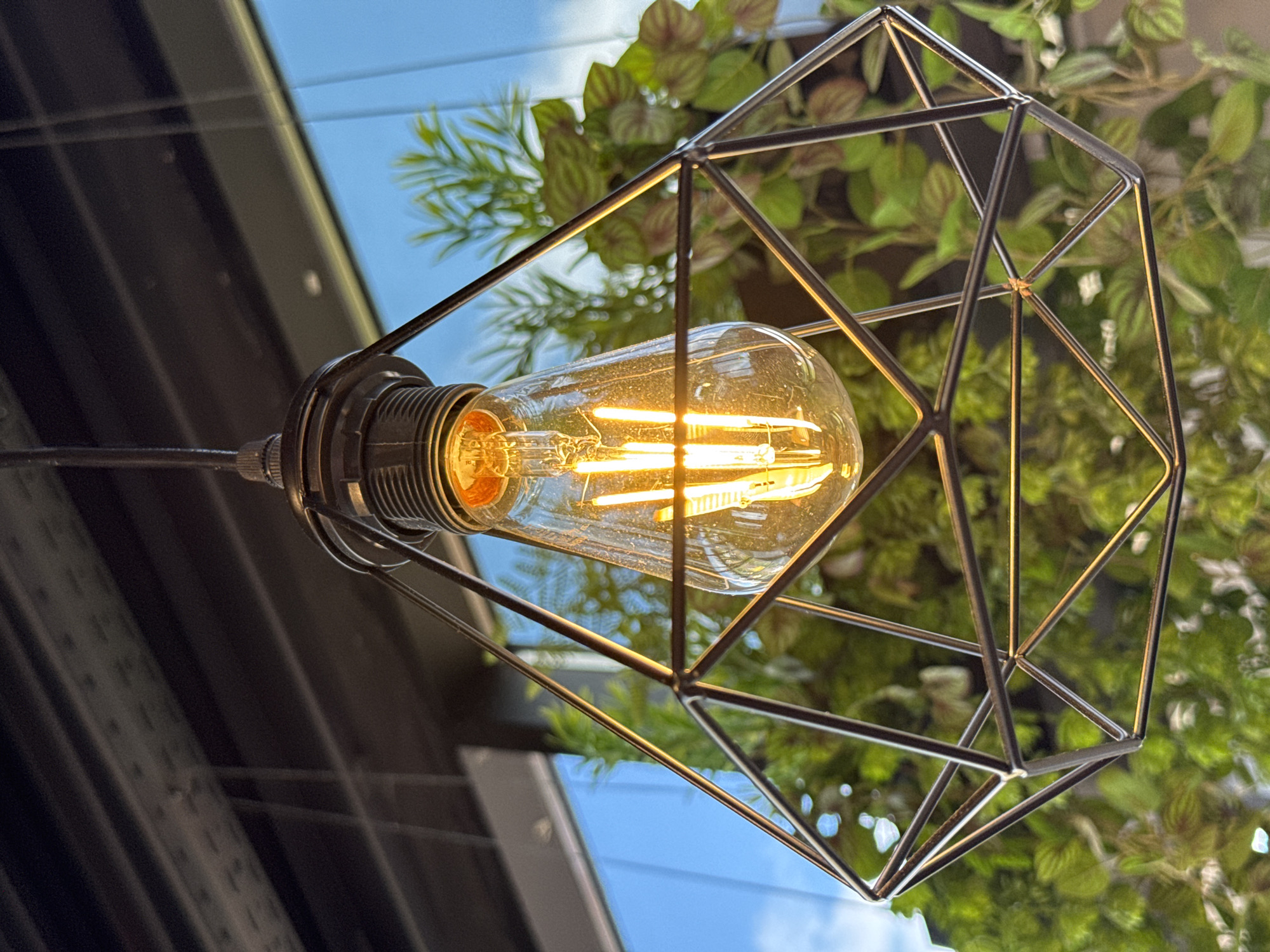 A close-up, low-angle shot captures a hanging Edison-style light bulb illuminated with a warm, yellow glow. The bulb is encased within a geometric, wireframe pendant light fixture, composed of intersecting black metal rods forming a faceted shape, resembling a diamond. The fixture is suspended from a dark ceiling by a thin cable. In the background, out of focus, is a large window with a bright blue sky and white clouds visible. Lush green foliage from indoor plants is also present, partially obscuring the window and casting shadows on the wireframe. The overall scene suggests an interior setting with natural light and decorative elements. There are no people or animals present. The location is specified as Peski, Russia.