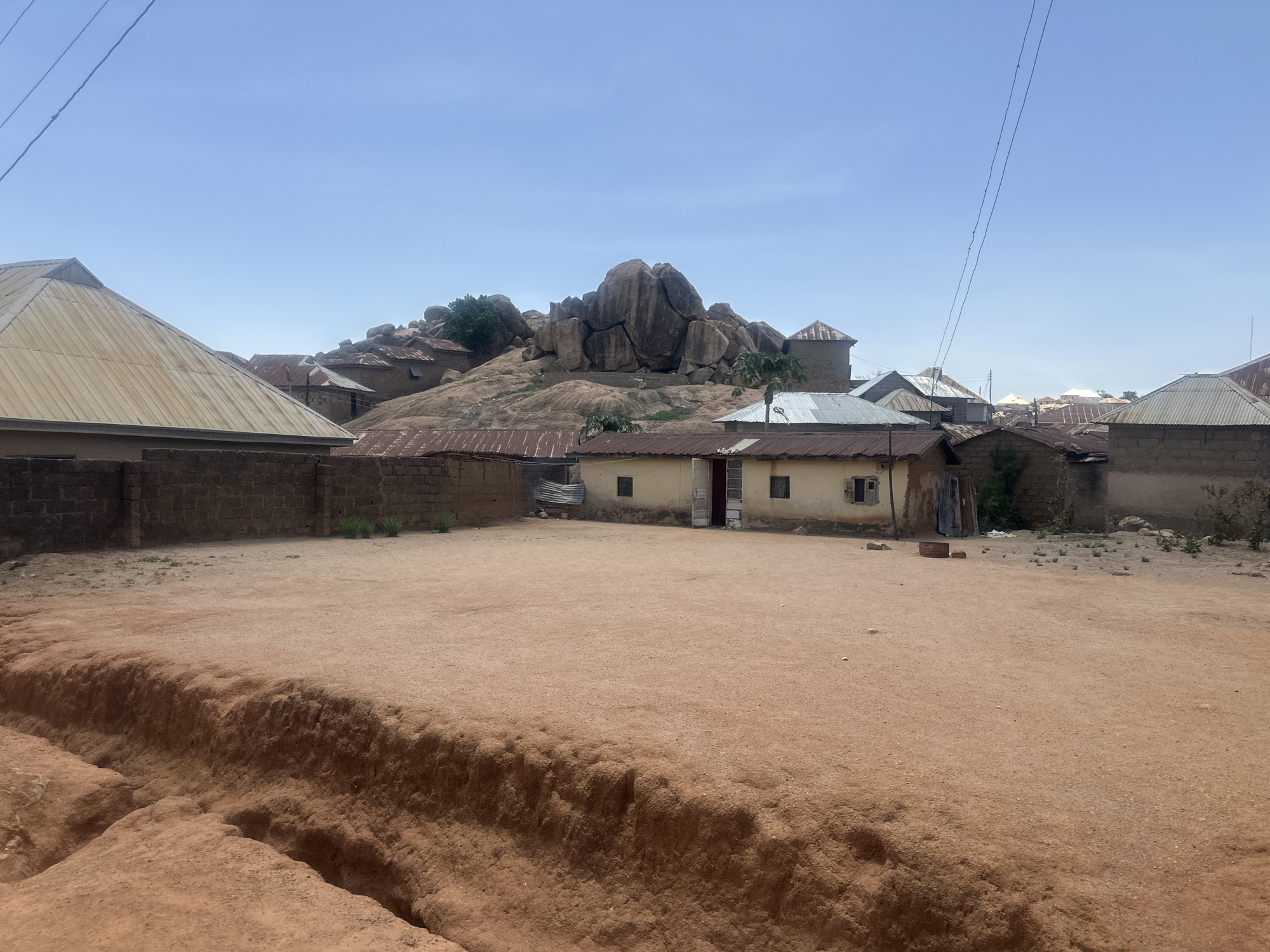 The scene depicts a dusty, open area in Jos, Nigeria, surrounded by buildings. In the background, a rocky outcrop with houses built near and around its base dominates the landscape. The buildings are constructed primarily of earth and brick, and many are topped with corrugated metal roofs. Power lines traverse the sky above the scene. The foreground consists of a dirt-covered area with a small gully. There are no visible people or animals in the frame.