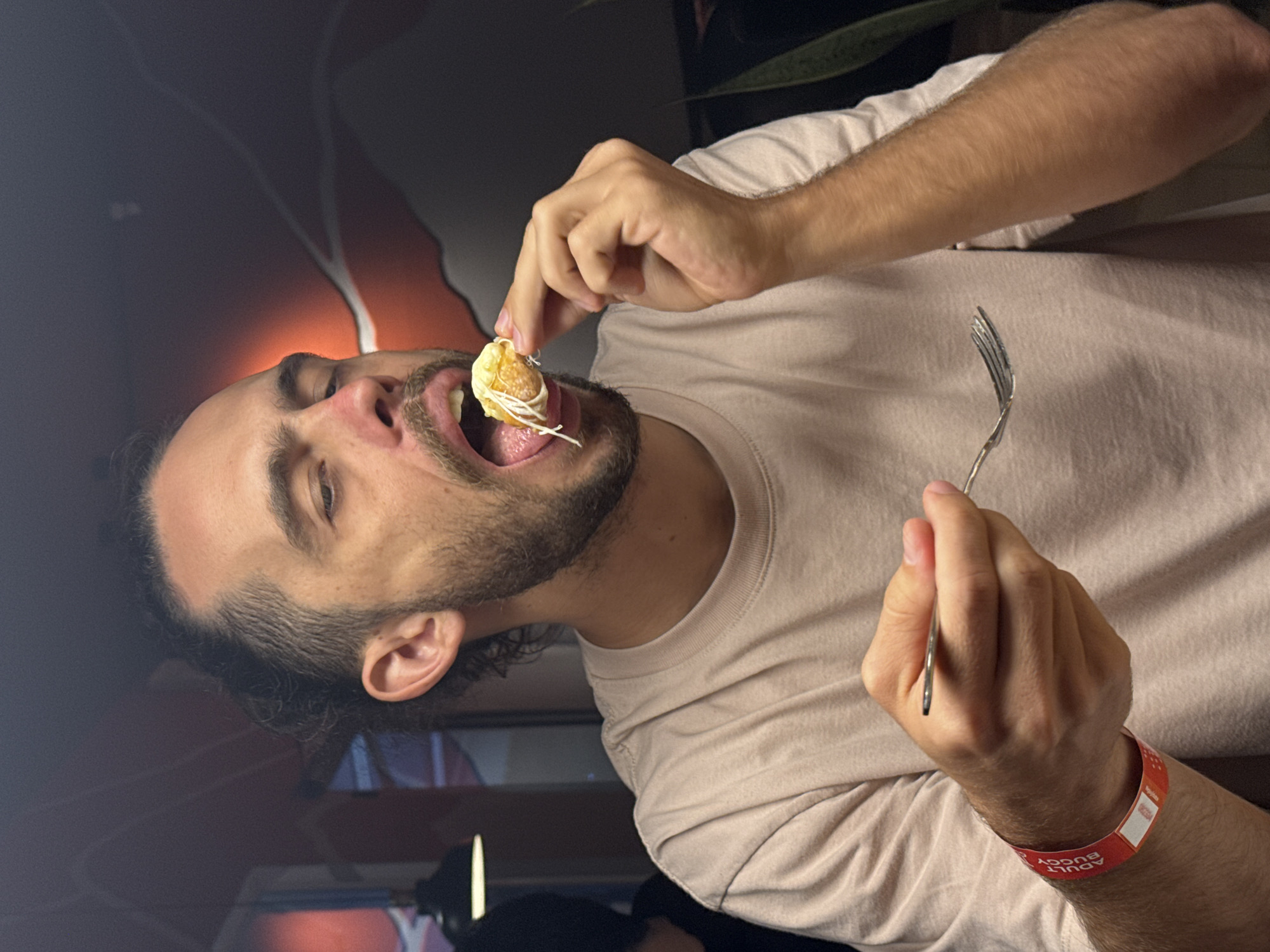 A man with dark hair and a beard is holding a piece of food close to his mouth with his right hand. He also holds a fork in his left hand. The man is wearing a light tan t-shirt and a red wristband that says "ADULT BUGGY". In the background, there is a dark wall and what appears to be a TV with a faint image of what could be people standing. The photo is indoors, and it is difficult to determine the exact location. There is no information in the image that indicates Đà Nẵng, Vietnam.