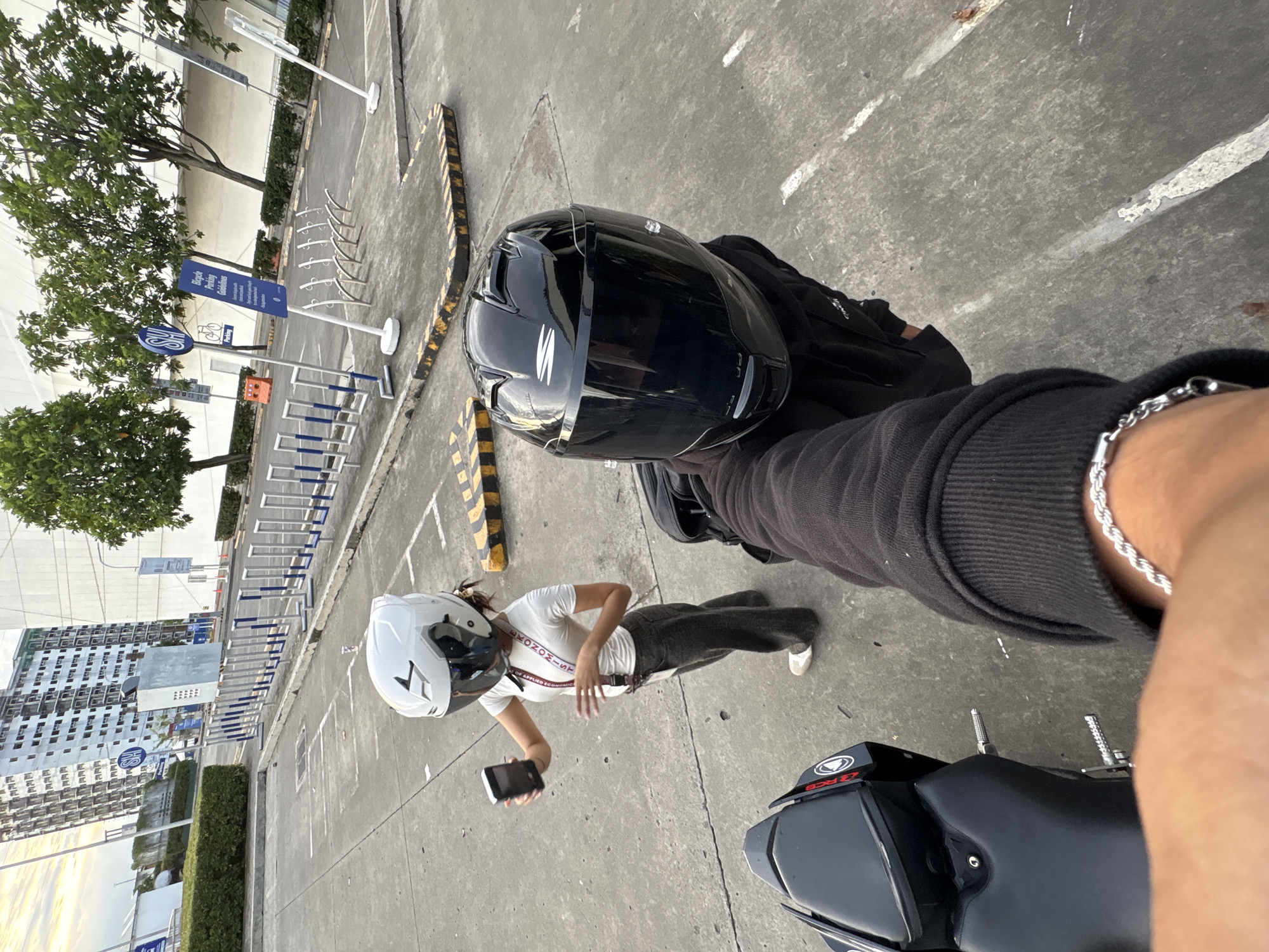 The image, taken in Davao City, Philippines, shows a top-down perspective from a motorcycle rider. The rider's arm, wearing a silver bracelet and a black long-sleeved shirt, is visible. A black motorcycle helmet rests on the seat in front of the rider. Standing nearby is a person wearing a white helmet, white t-shirt, dark jeans, and white shoes. They are holding a smartphone. The motorcycle, with the "RCB" label visible, is parked on a concrete surface. In the background, there is a bicycle parking area, a building, trees, and street markings.