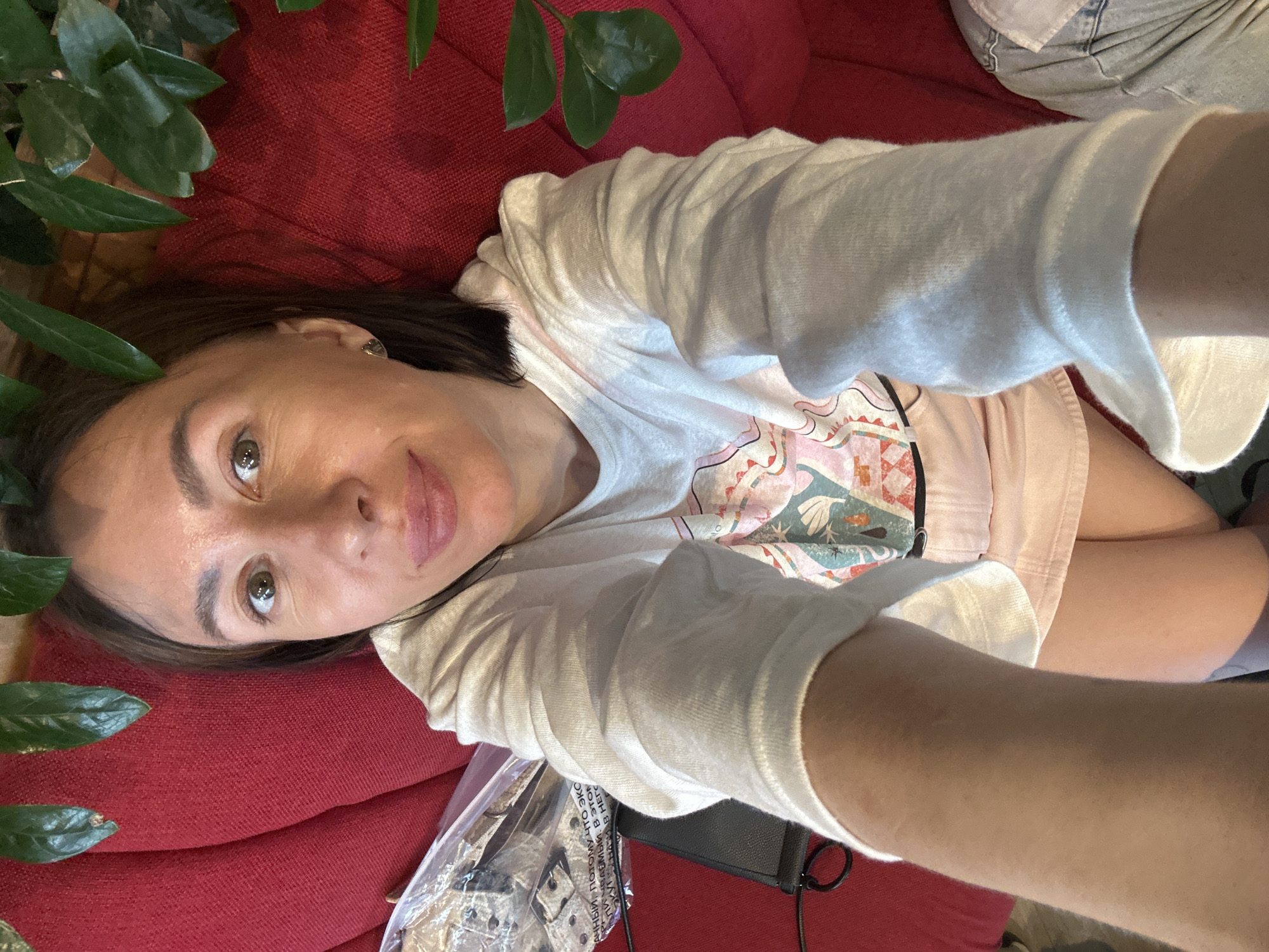 An overhead, medium shot captures a woman lying on a red sofa, possibly in a residential setting in Izhevsk, Russia. She is wearing a white long-sleeved shirt with a colorful graphic print and a light pink skirt. Green leaves from an overhead plant frame the top of the image. To her left, a black handbag and a clear plastic bag containing what appears to be clothing or fabric are visible on the sofa. Her arms are extended towards the camera, as if taking a selfie. The overall lighting is soft and natural.