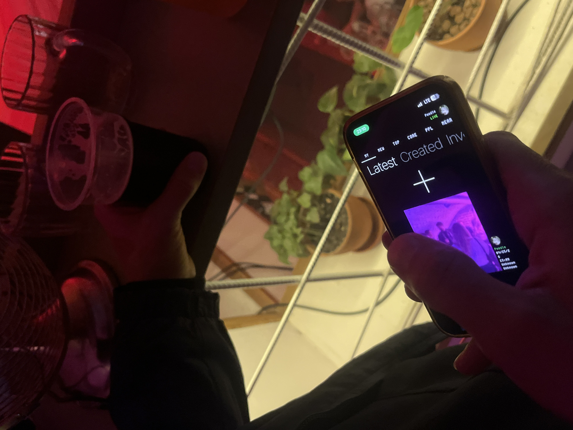 A person is holding a smartphone with a screen displaying the word "Latest Created Inv" along with a plus sign and other text. The phone screen also shows the time as "23:10". The person is wearing dark clothing, and the background reveals glimpses of houseplants, shelves, and what appears to be an electric fan. The time on the phone is 23:10 which suggests this image may have been taken somewhere where they use a 24 hour clock. It is impossible to know where this image was taken without additional information.