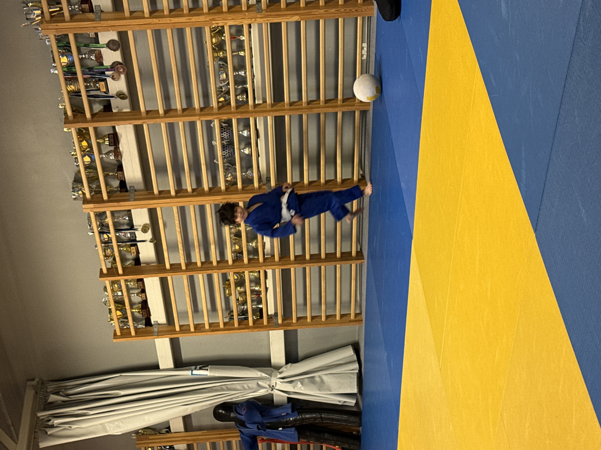 A young boy in a blue judo uniform with a white belt is running on a blue and yellow mat, kicking a white ball. He is in a gymnasium with wooden wall bars and shelves filled with trophies and medals. To the left of the boy is a dark blue training dummy wearing a blue judo uniform. A light gray curtain is visible on the far left of the image. The boy appears to be in an indoor sports facility in Tallinn, Estonia.