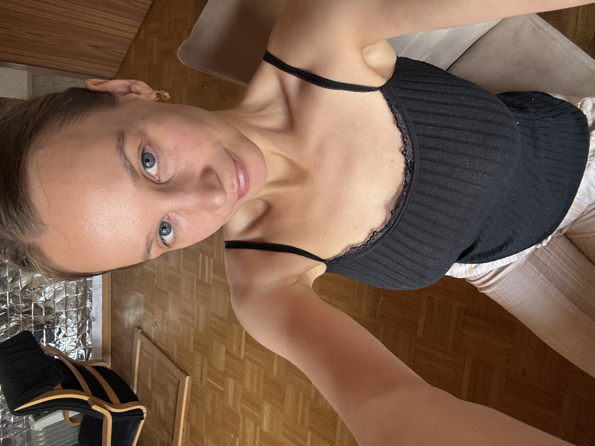 A young woman with blue eyes and light brown hair pulled back is captured in a close-up selfie. She is wearing a black tank top with lace trim and light-colored pants. She is seated on a chair in what appears to be a room with wooden flooring and a chair in the background. The overall lighting suggests an indoor setting, possibly a home. The image does not provide any specific information to place it in Überherrn, Germany.