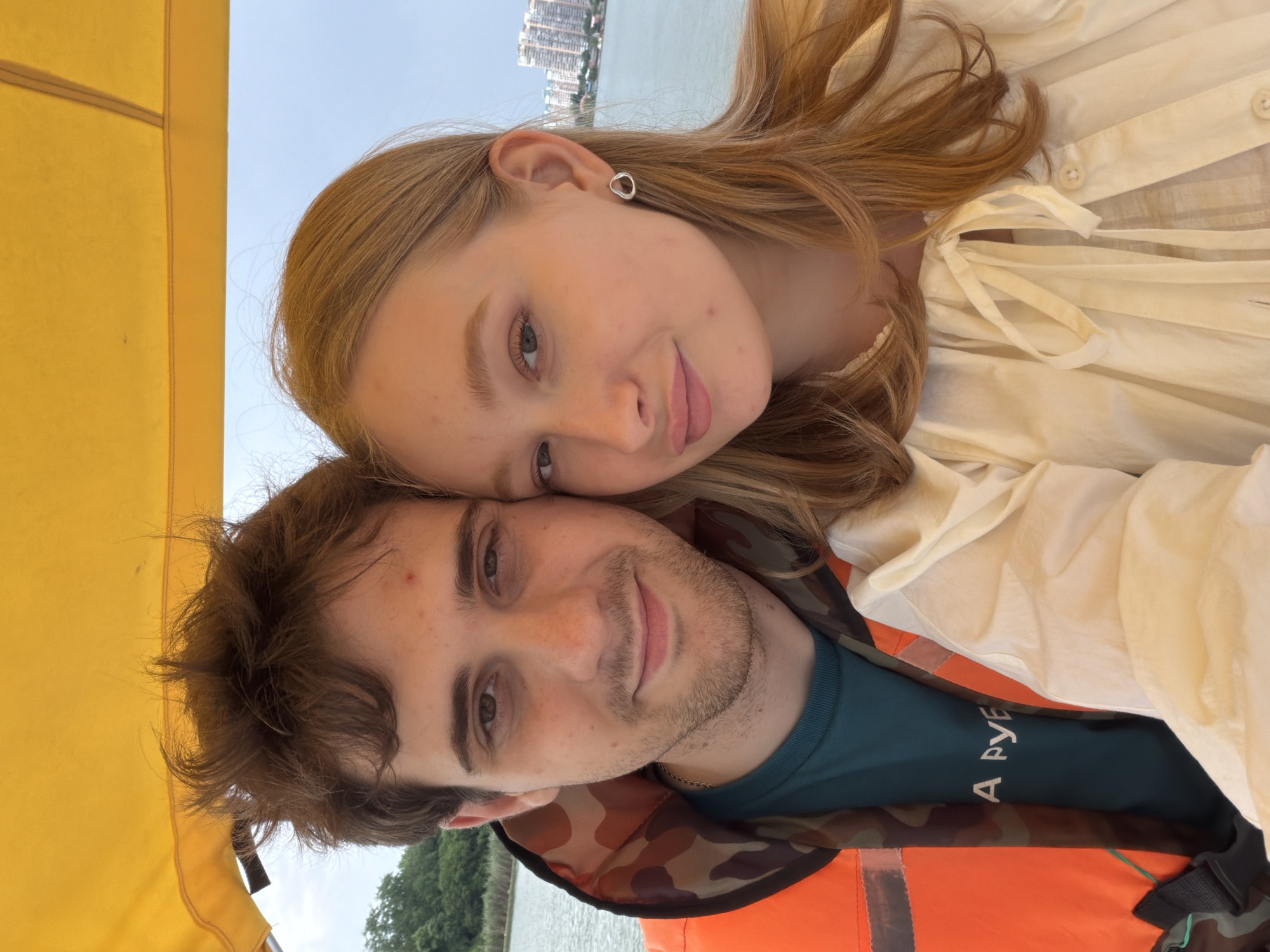 A selfie taken from a slightly high angle shows a young couple on a boat with a yellow canopy. The man, on the left, has brown hair and is wearing an orange life vest over a teal shirt. The woman, on the right, has blonde hair and is wearing a white top. They are both looking at the camera and smiling. The background shows a body of water, trees on the left bank, and buildings on the right bank, suggesting they are on a river or a large canal within the city of Krasnodar, Russia.